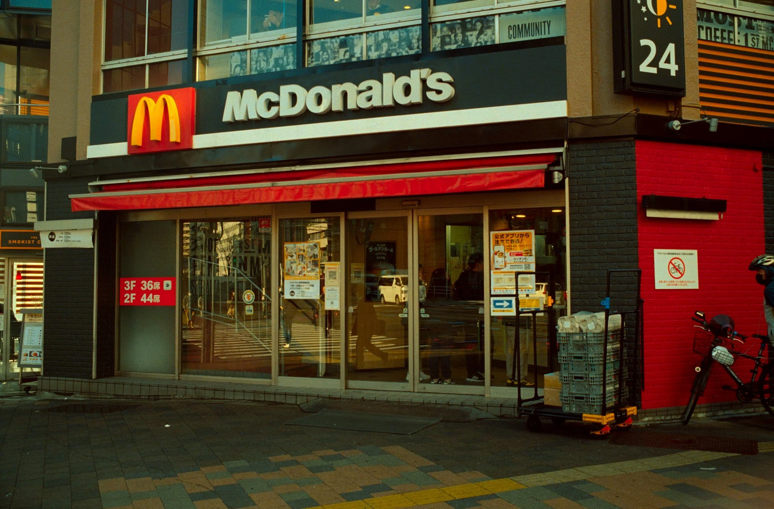 Exterior view of a McDonald's restaurant in an urban area, with a black and red facade, glass windows, and a sign showing the store hours. There are bicycles parked outside and some signage with Japanese text.