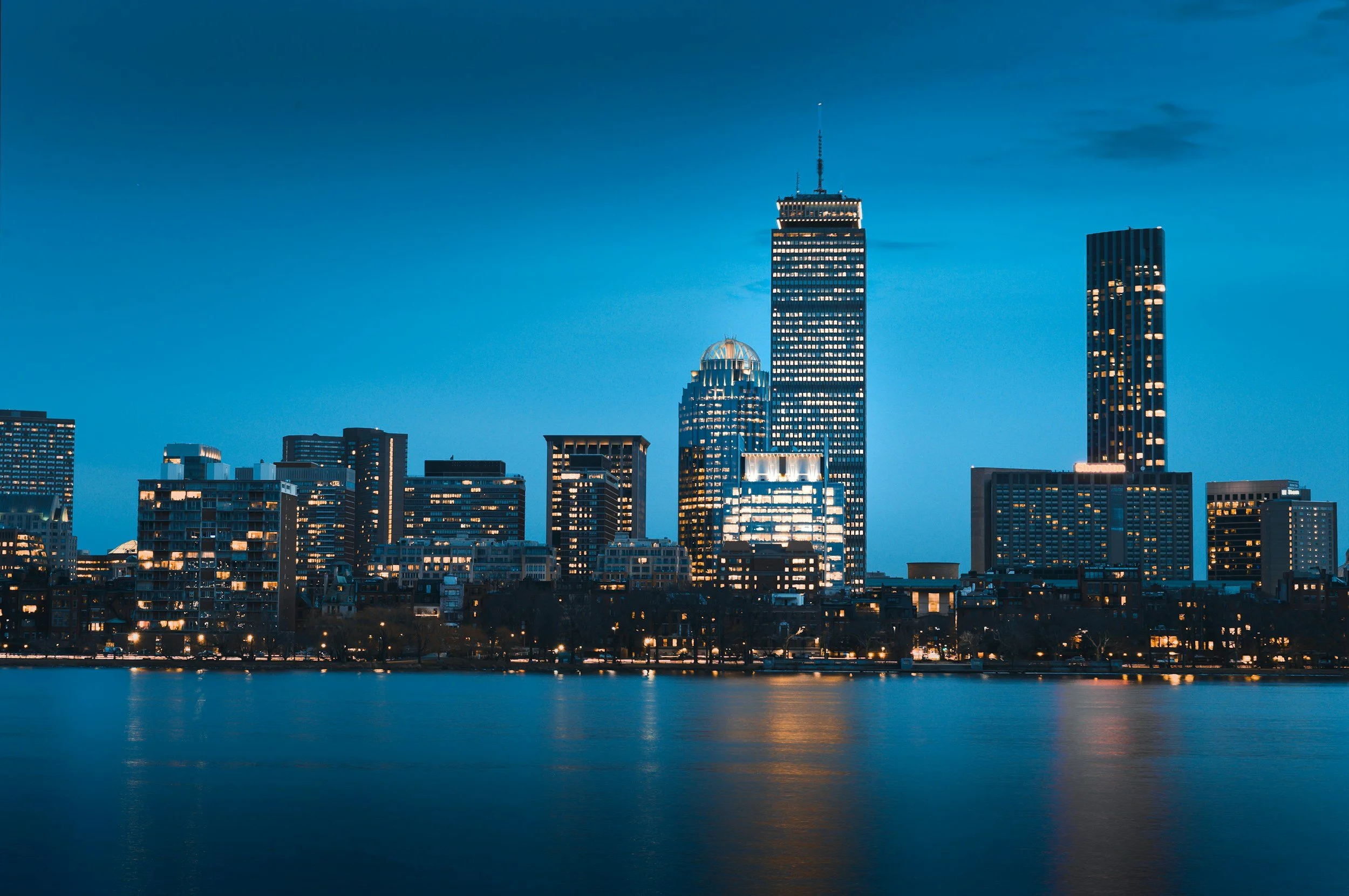 Nighttime city skyline with illuminated skyscrapers reflected in a body of water.