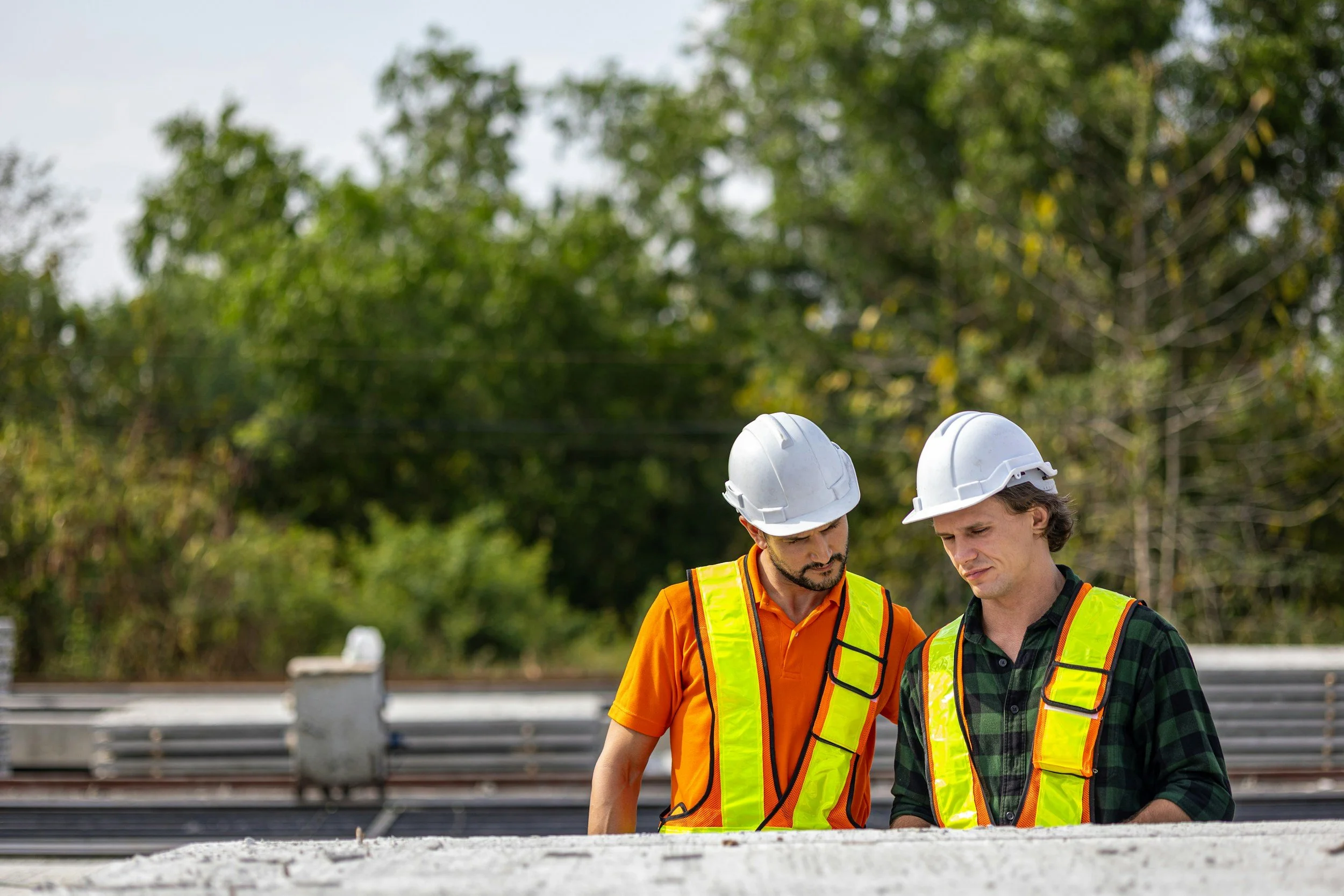 Two construction workers wearing safety helmets and vests looking at plans outdoors with trees in the background.
