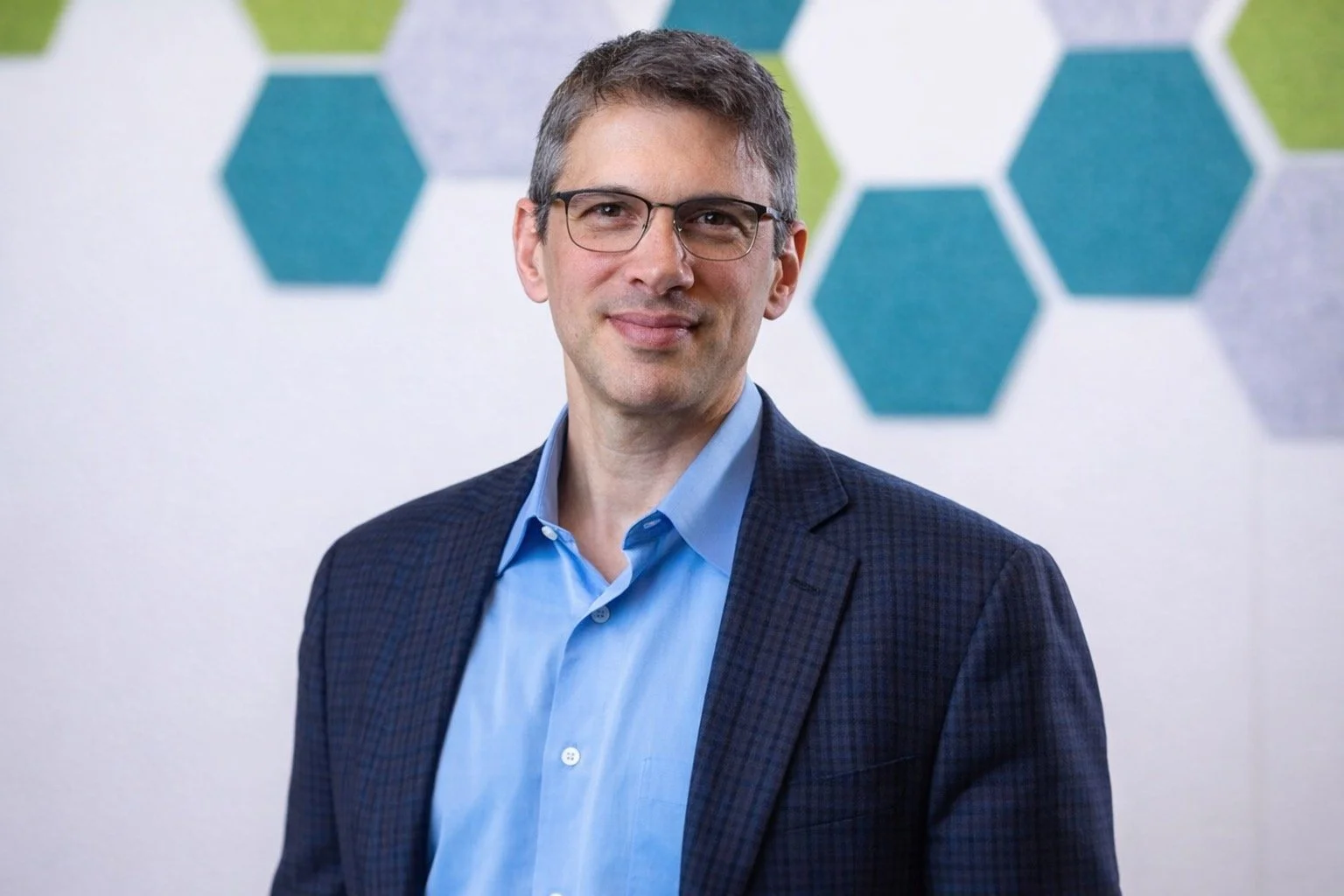 A man with short brown hair and glasses, wearing a blue dress shirt and dark blazer, standing in front of a wall with a pattern of hexagons in shades of blue and green.