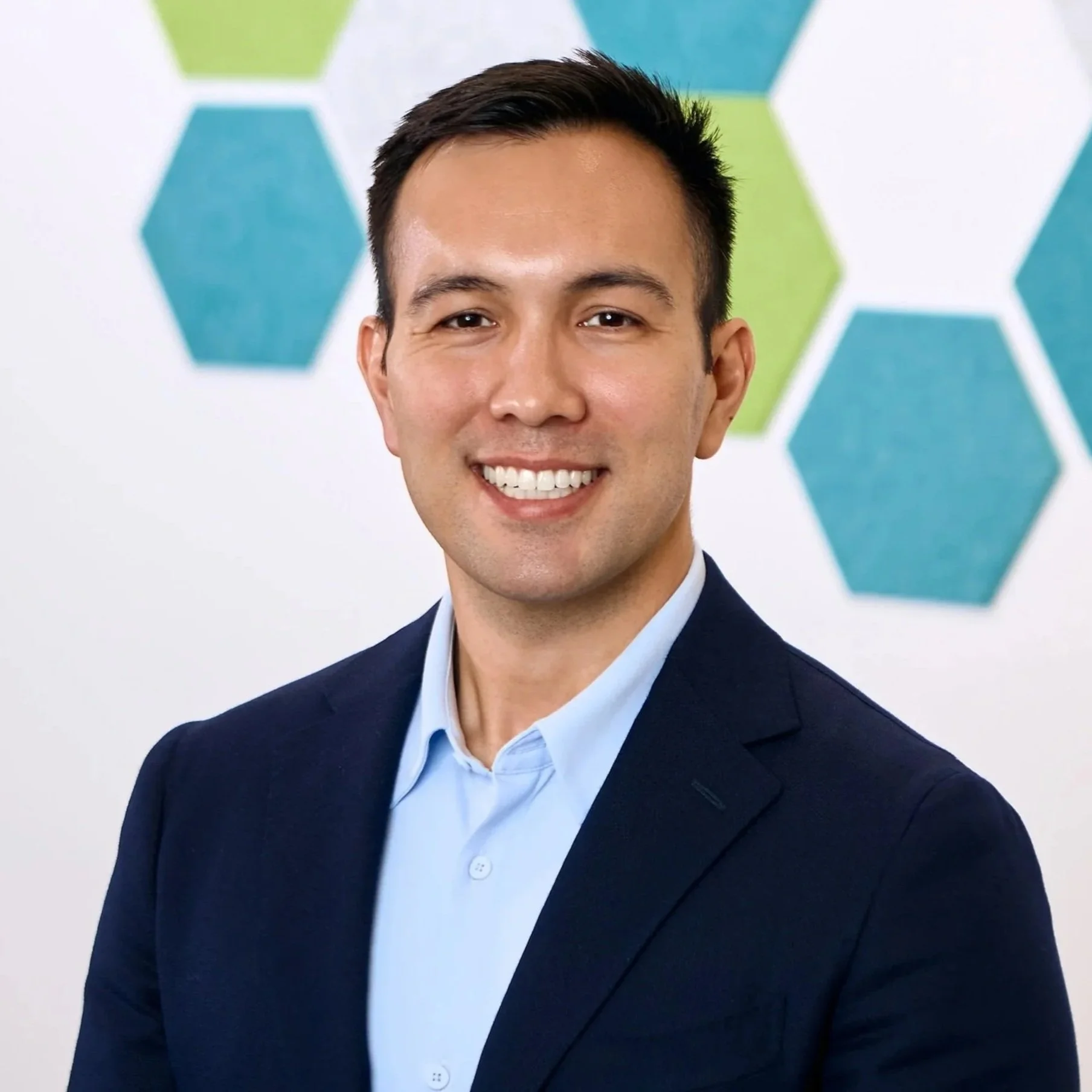 A professional man smiling, wearing a navy blazer and a light blue shirt, with a background featuring hexagon shapes in shades of blue and green.