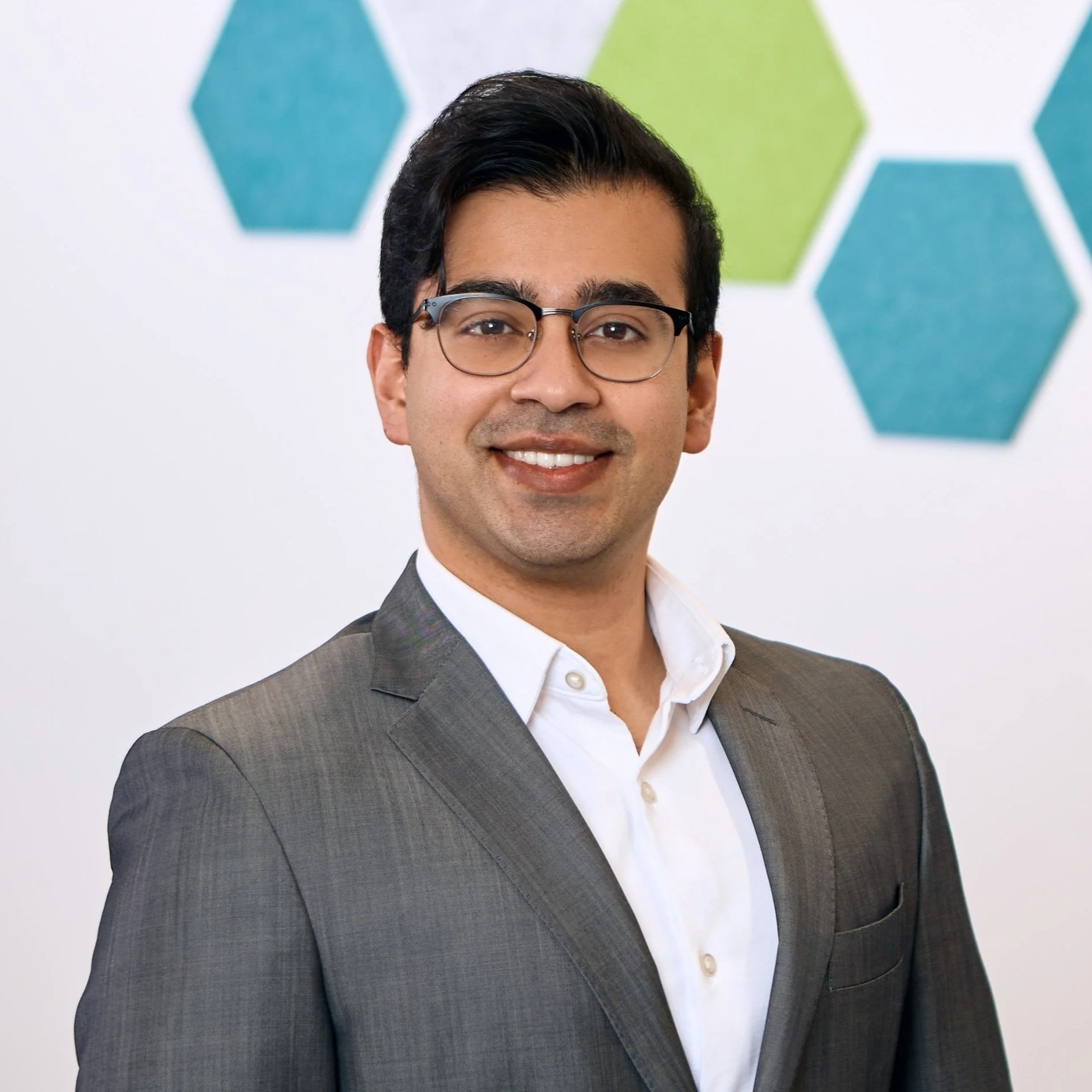 A man wearing a gray business suit and glasses, smiling, with a white shirt, standing in front of a white wall decorated with green and blue hexagon shapes.