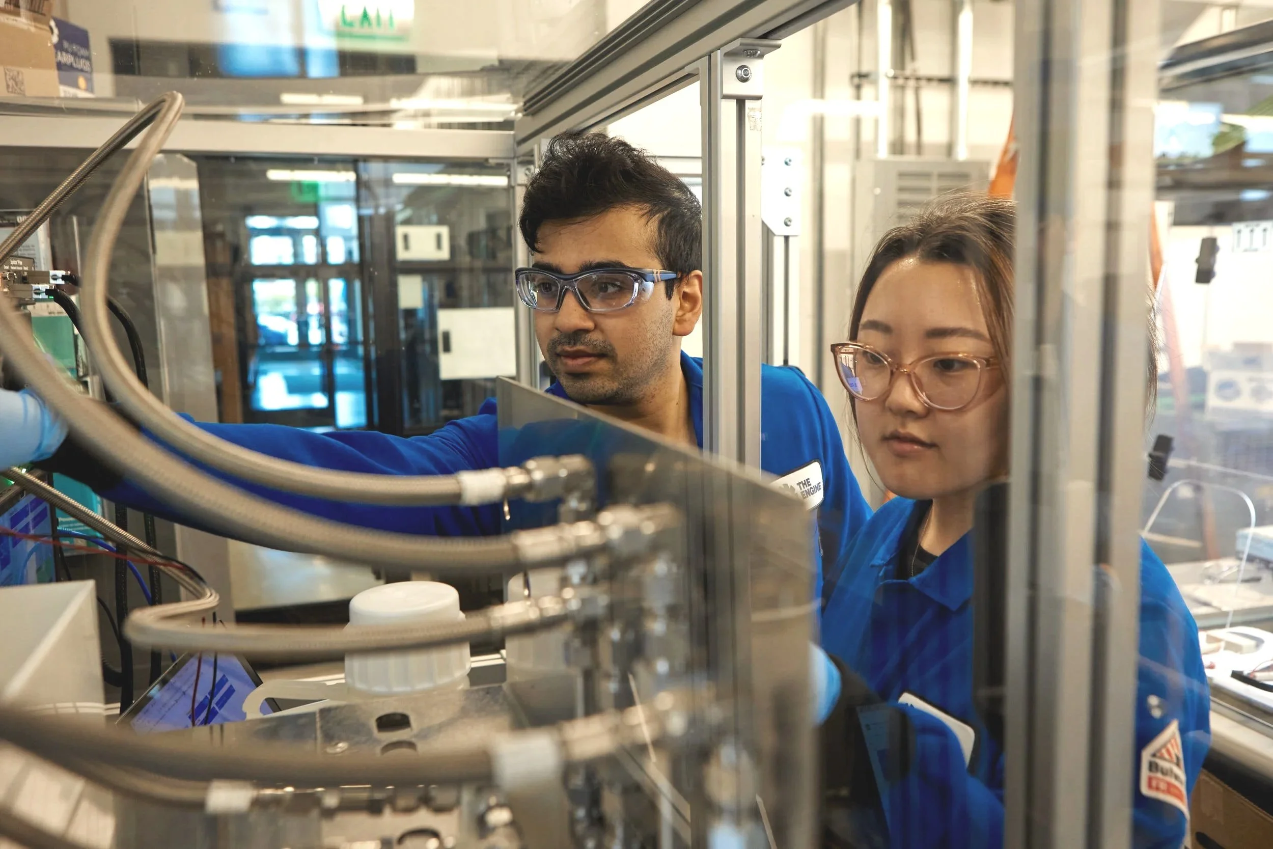 Two scientists working in a laboratory, wearing blue lab coats and safety glasses, inspecting equipment behind a glass enclosure.