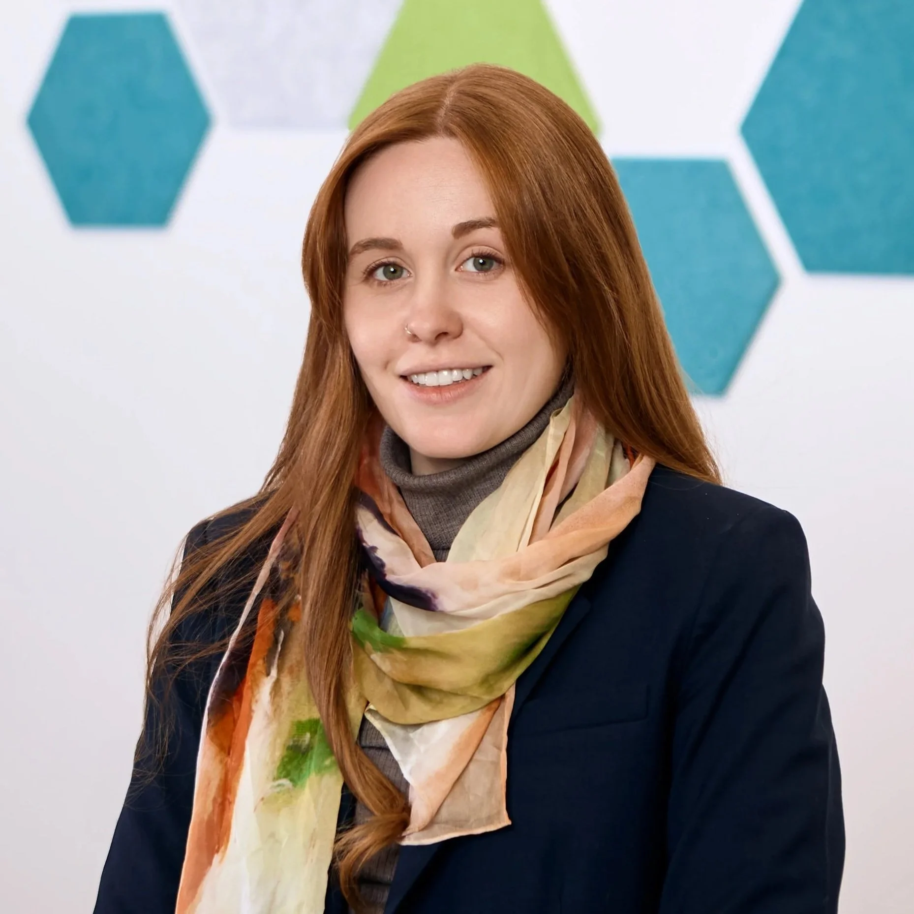 A woman with long red hair, wearing a multicolored scarf, dark blazer, and a gray turtleneck, smiling in front of a background with blue and green hexagon shapes.