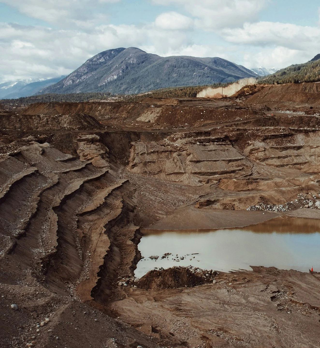 A landscape with a large open-pit mining quarry, showing terraced earth walls, a body of water at the bottom, and mountains in the background under partly cloudy skies.