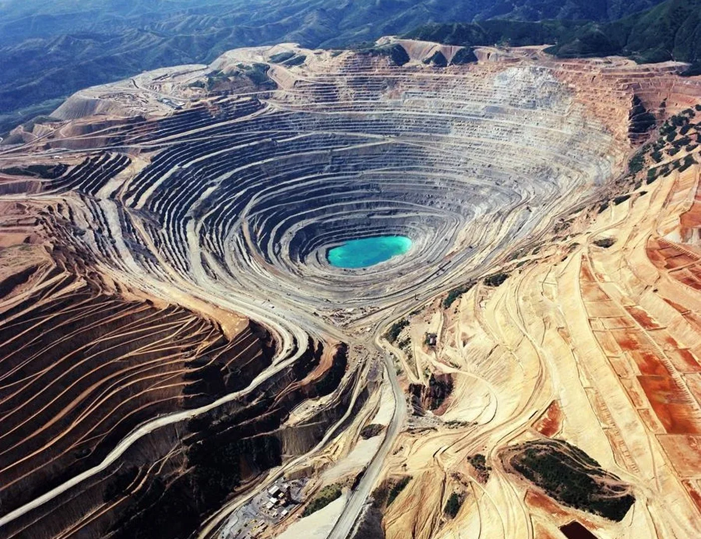 An aerial view of a large open-pit copper mine with terraced levels surrounding a turquoise pit lake at its center, with surrounding hills and mountains in the background.