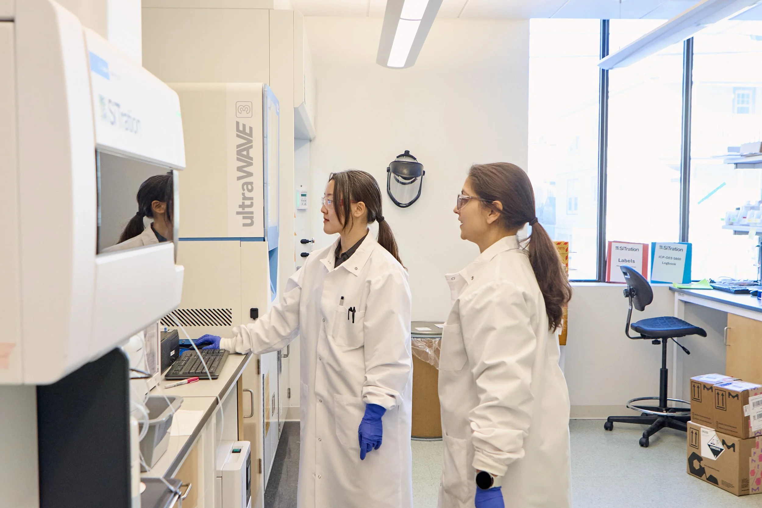 Two women scientists in white lab coats and safety glasses working in a laboratory with scientific equipment, boxes, and large windows in the background.