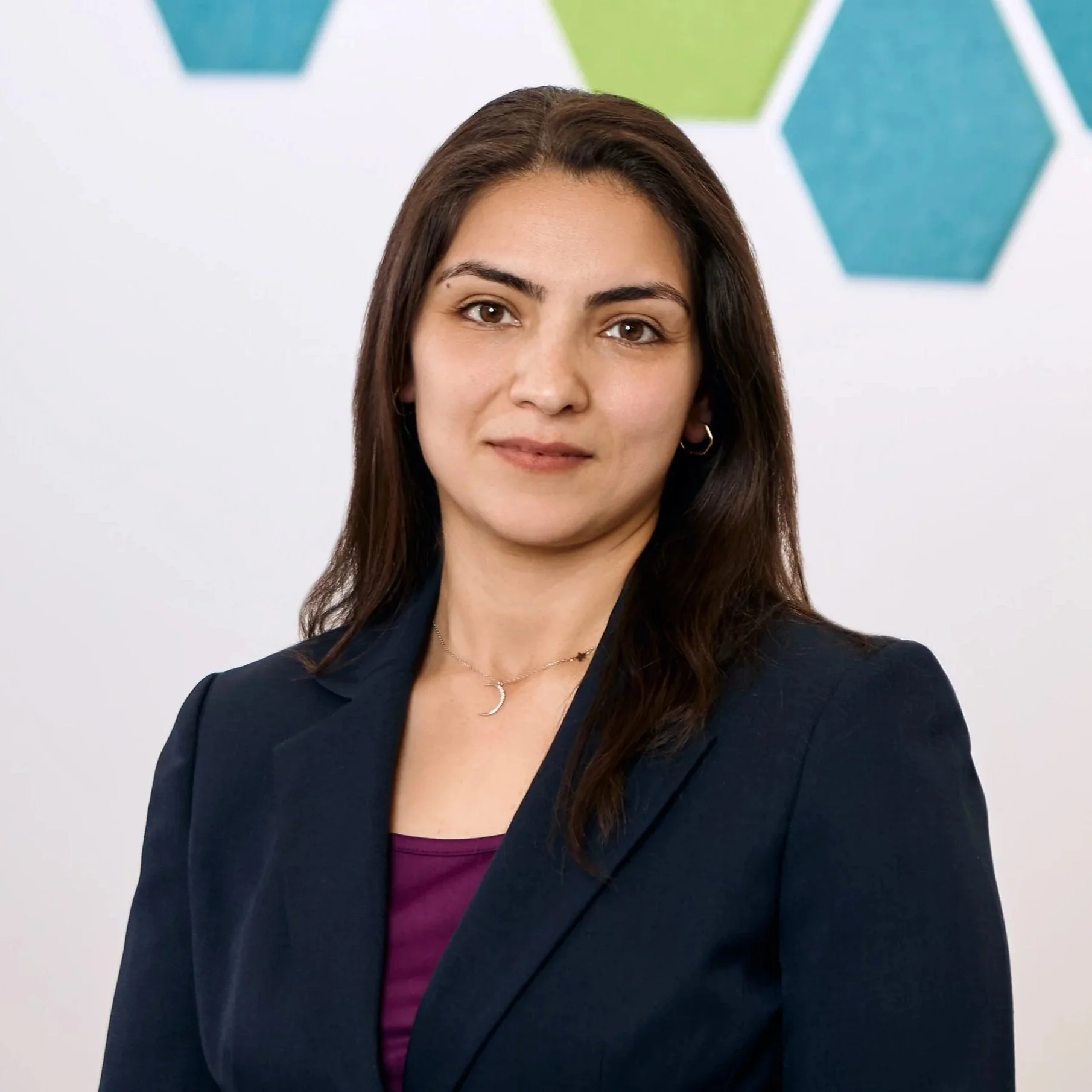 A woman with long dark hair wearing a navy blazer and purple top, standing against a white background with colorful hexagons.