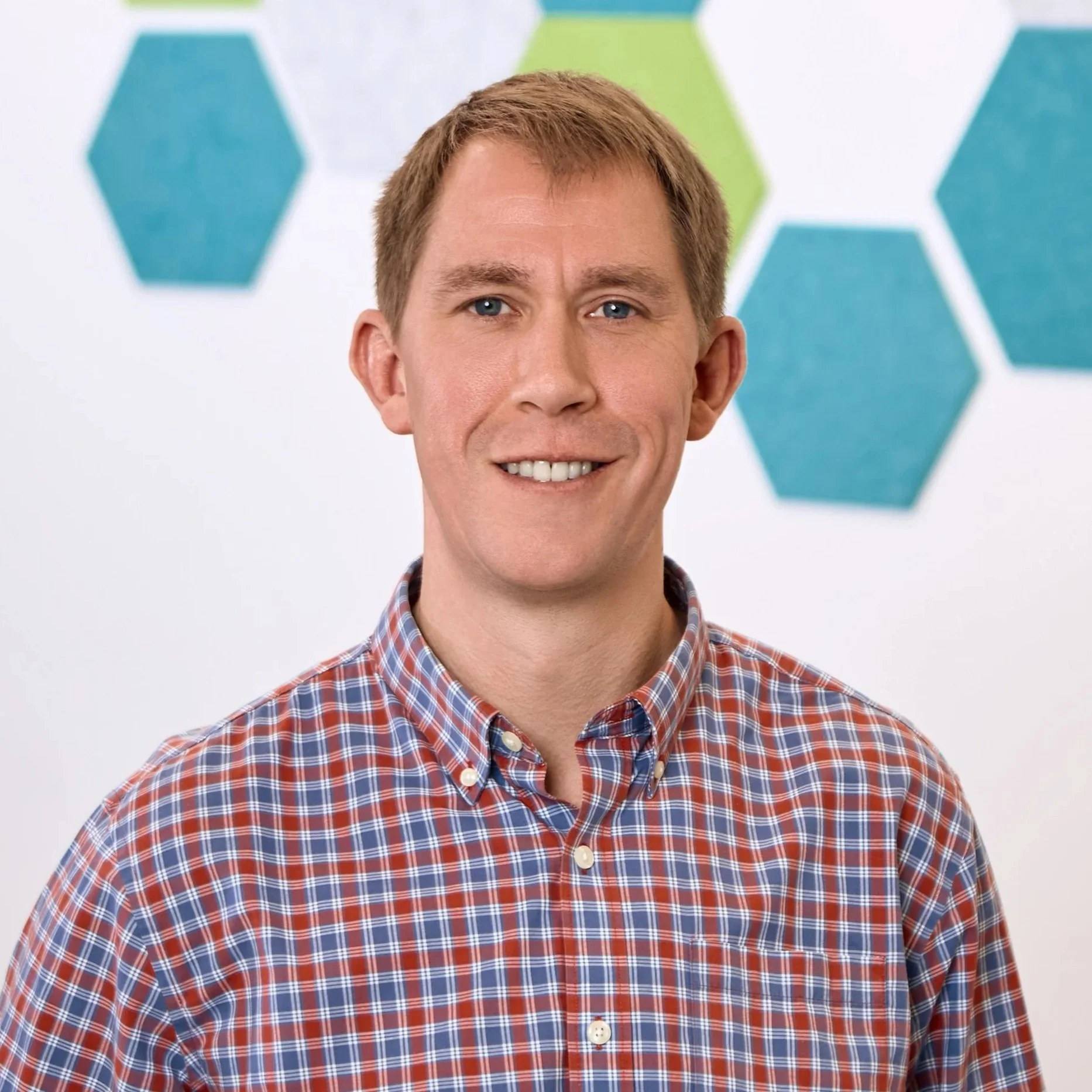 Portrait of a smiling man with short, light brown hair, wearing a red and blue checked shirt, standing in front of a white background with colorful hexagon patterns.