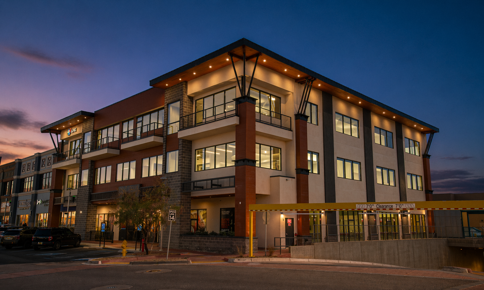Modern multi-story building with large windows and balconies at dusk, with a parking lot in the foreground and a clear sky with sunset colors in the background.