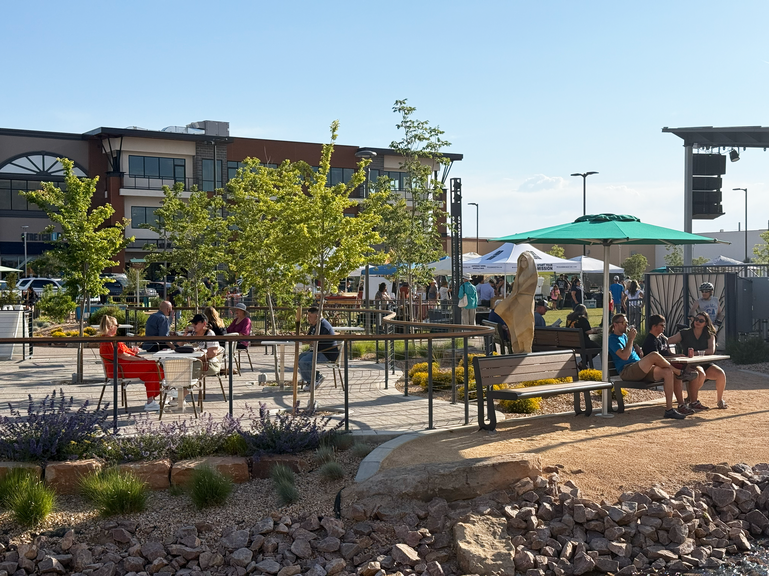 People sitting and chatting at outdoor tables and benches in Winrock park with trees, plants, and a sunny sky.