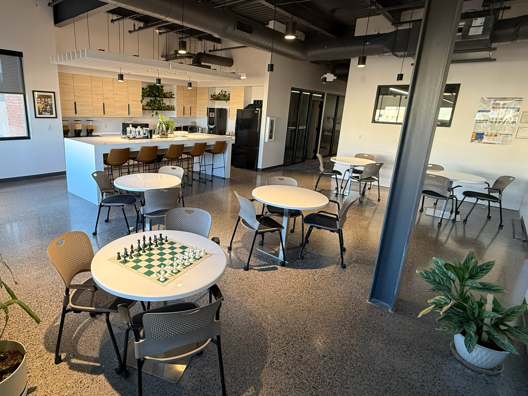 Modern coworking and kitchen common area with round tables and chairs. The background features a kitchen with a breakfast bar, brown stools, and shelves with plants.
