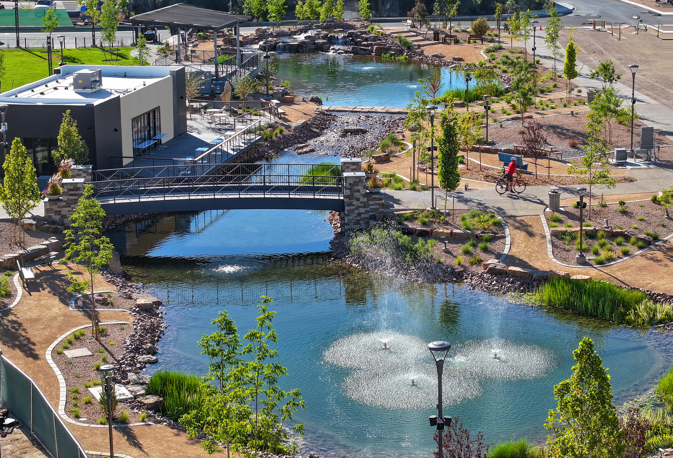 Bird's eye view of Winrock park with a winding pond, a small waterfall, a bridge, walking paths, trees, benches, and a person riding a bicycle.
