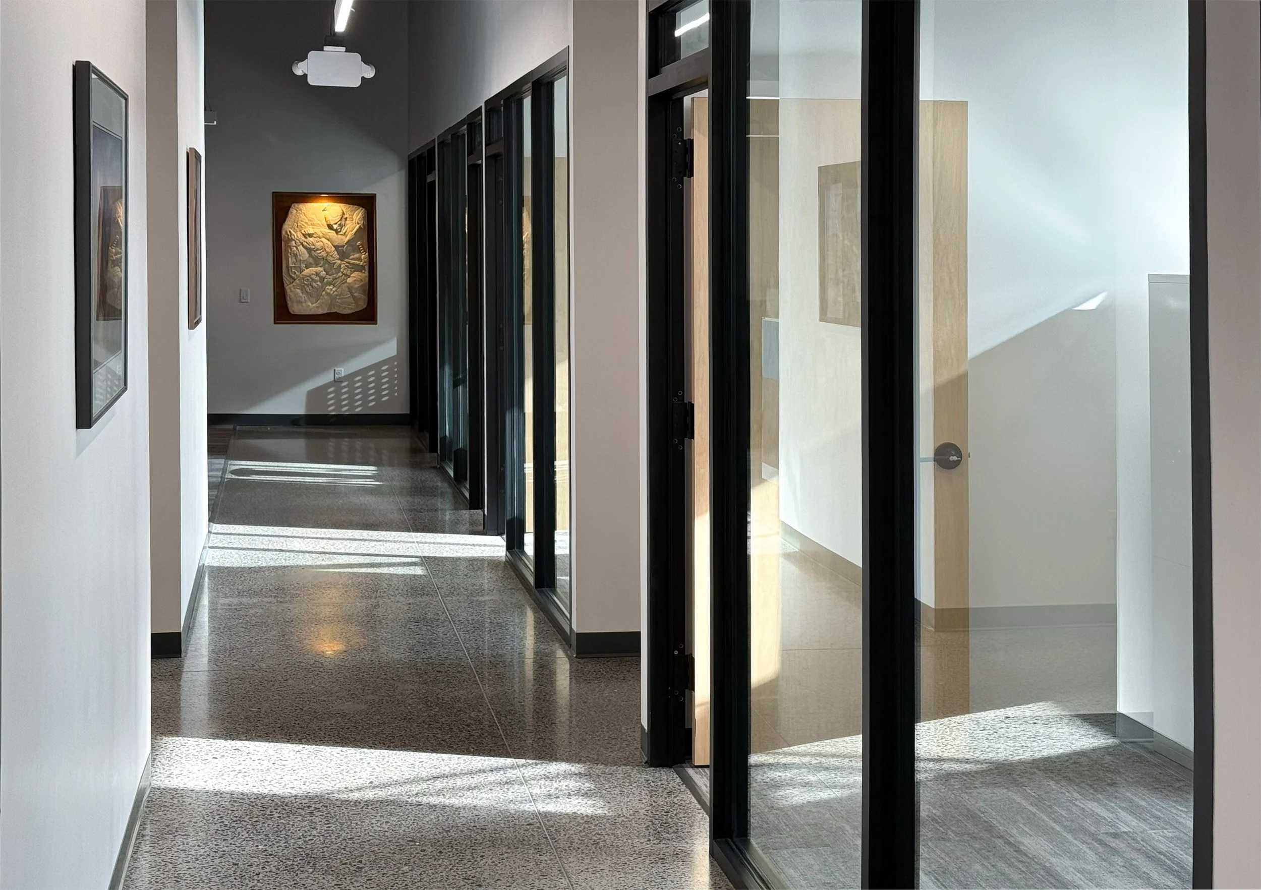 Modern office hallway with natural light, artwork on walls, glass partitions, and polished flooring.