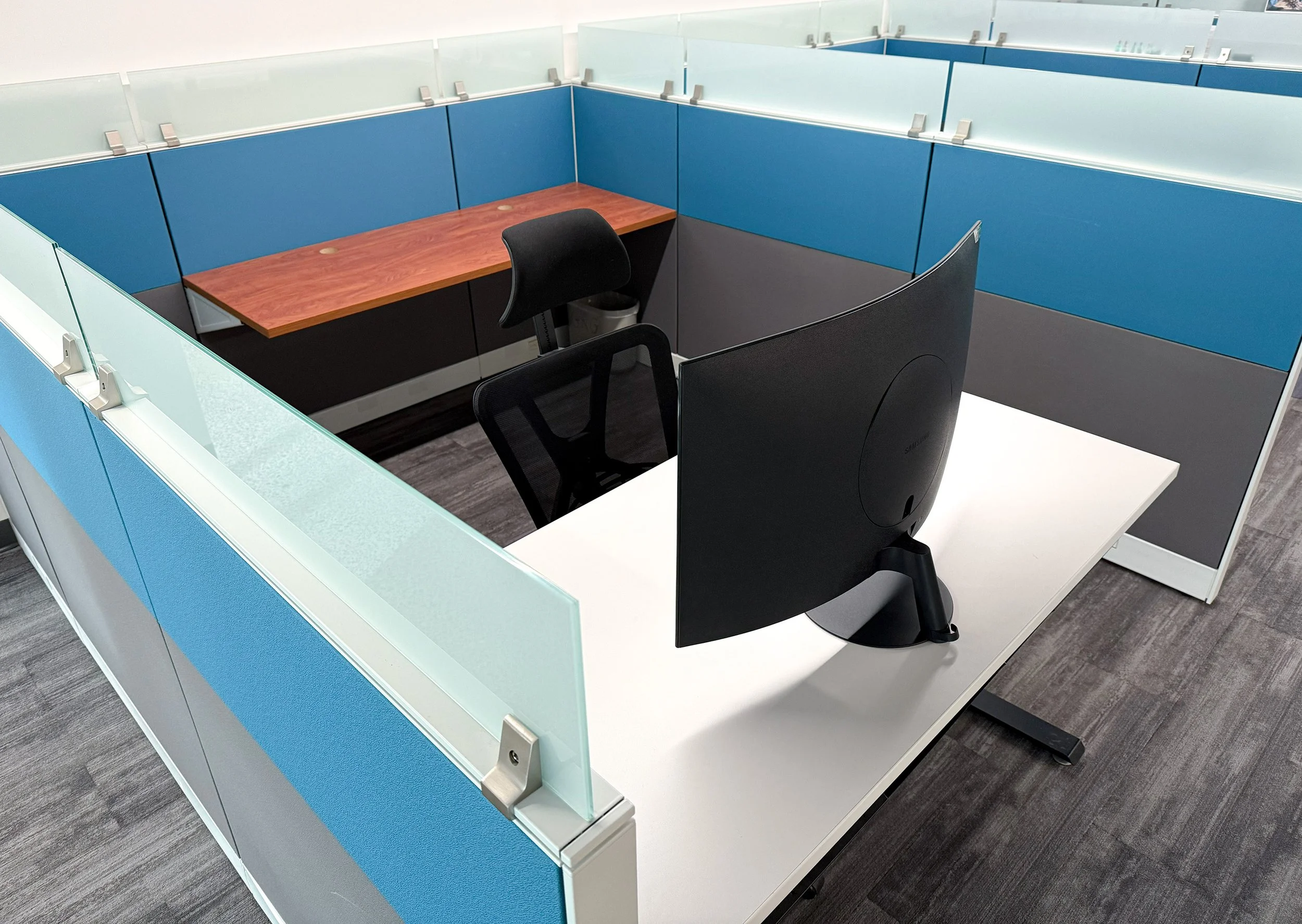 Move-in ready office cubicle with blue partitions, a wooden desk, a black office chair, and a computer monitor.