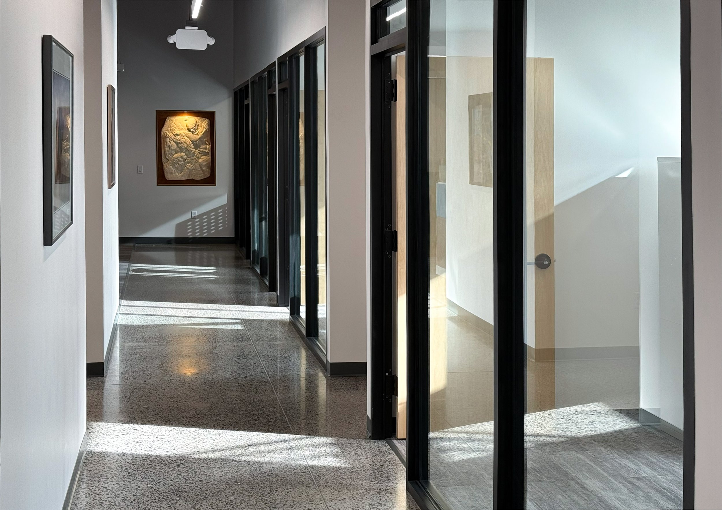 Modern office hallway with glass-walled conference rooms on the right and framed artwork on the left wall. Sunlight creates patterns on the polished floor.