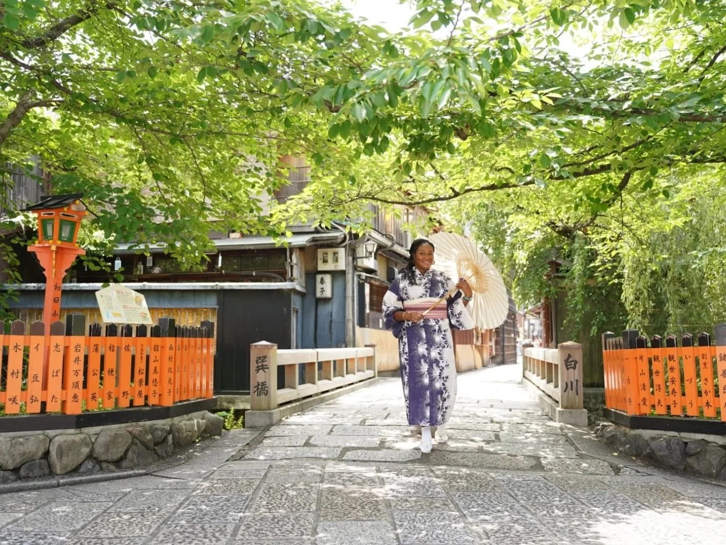 A woman wearing a traditional Japanese yukata walking on a stone-paved street, holding a parasol, surrounded by lush green trees and a fence with orange and black wooden plaques.