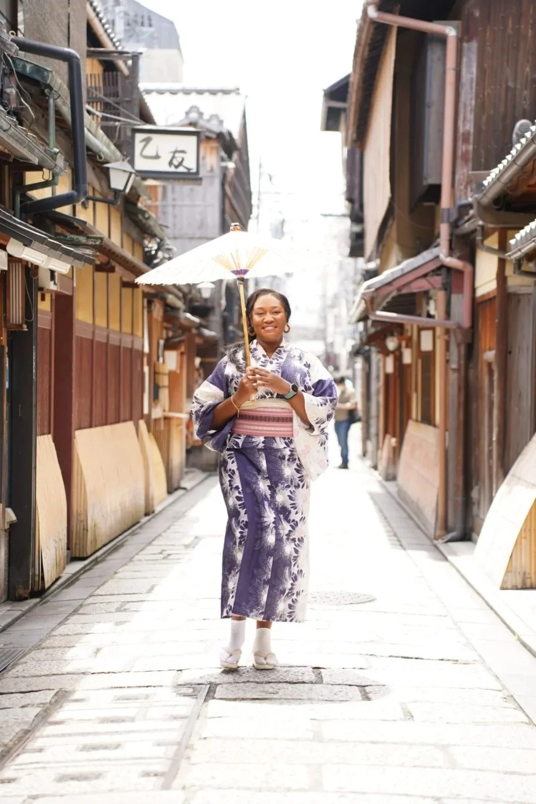 A woman wearing a kimono and holding a paper parasol standing in a narrow street with traditional wooden buildings.