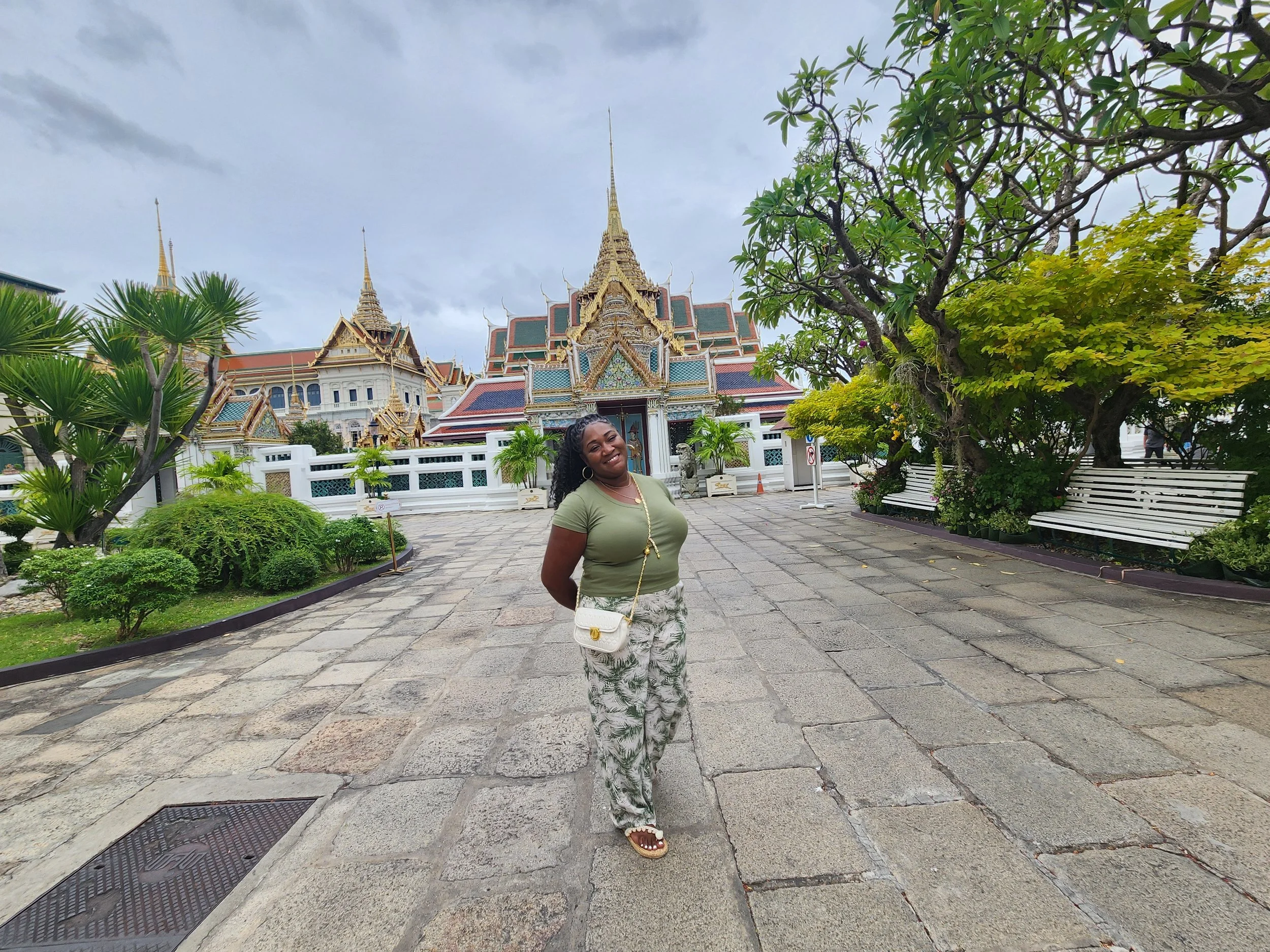 A smiling woman standing on a stone-paved walkway in front of a traditional Thai temple, with lush green trees and benches around.