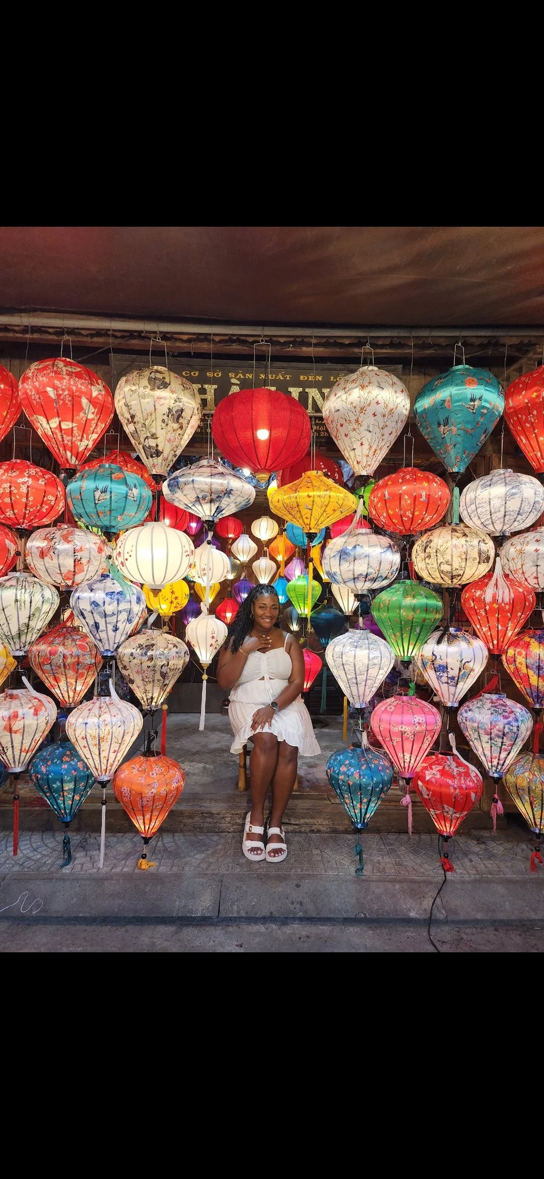 A woman in a white dress sitting on a bench in front of a vibrant display of colorful hanging lanterns.