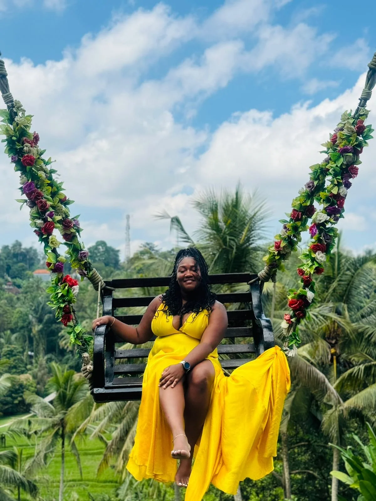 A woman in a yellow dress sitting on a decorated swing surrounded by green tropical trees and plants.