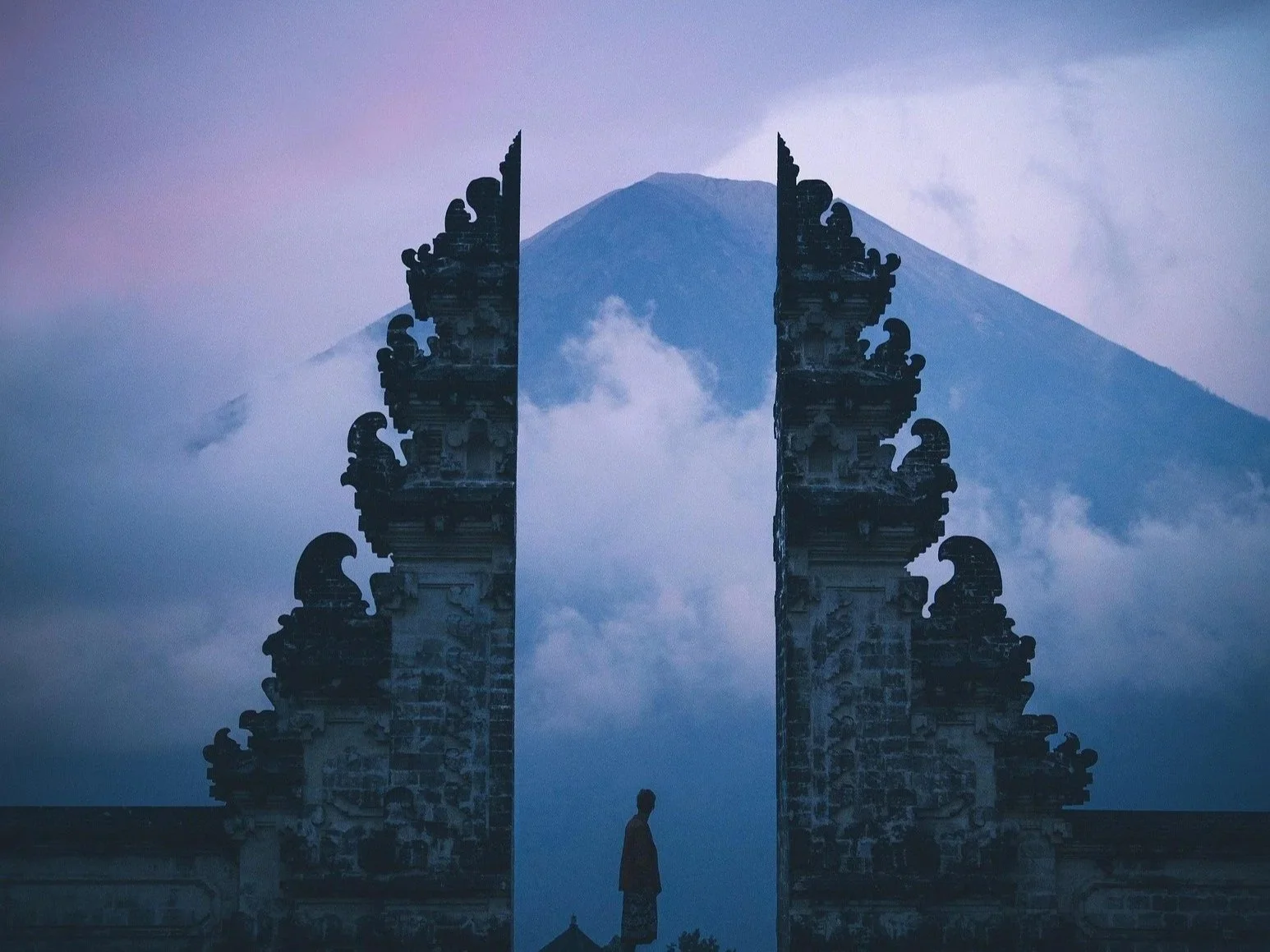 A traditional Balinese entrance gate with intricate carvings, silhouetted against a large mountain, possibly Mount Agung, in the background. There is a solitary person standing in the middle of the gate.