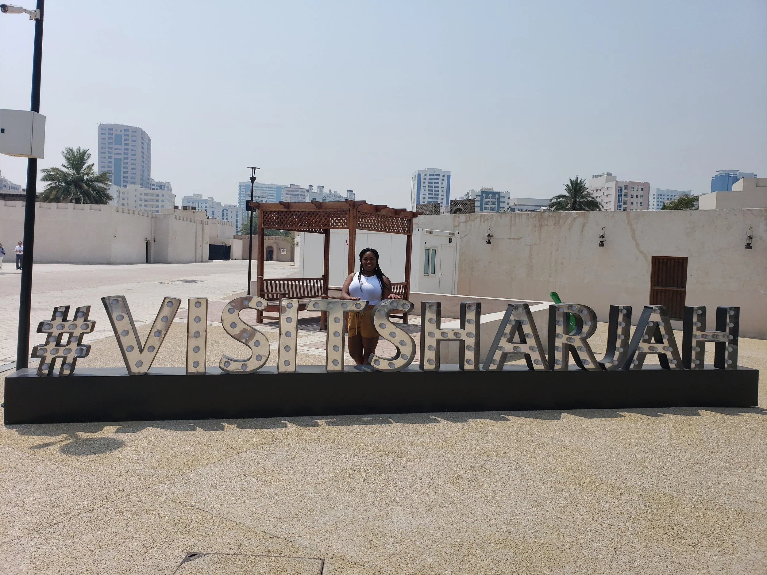 Woman standing behind a sign that reads '#VISITSHARJAH' in front of a beige wall with city buildings in the background.