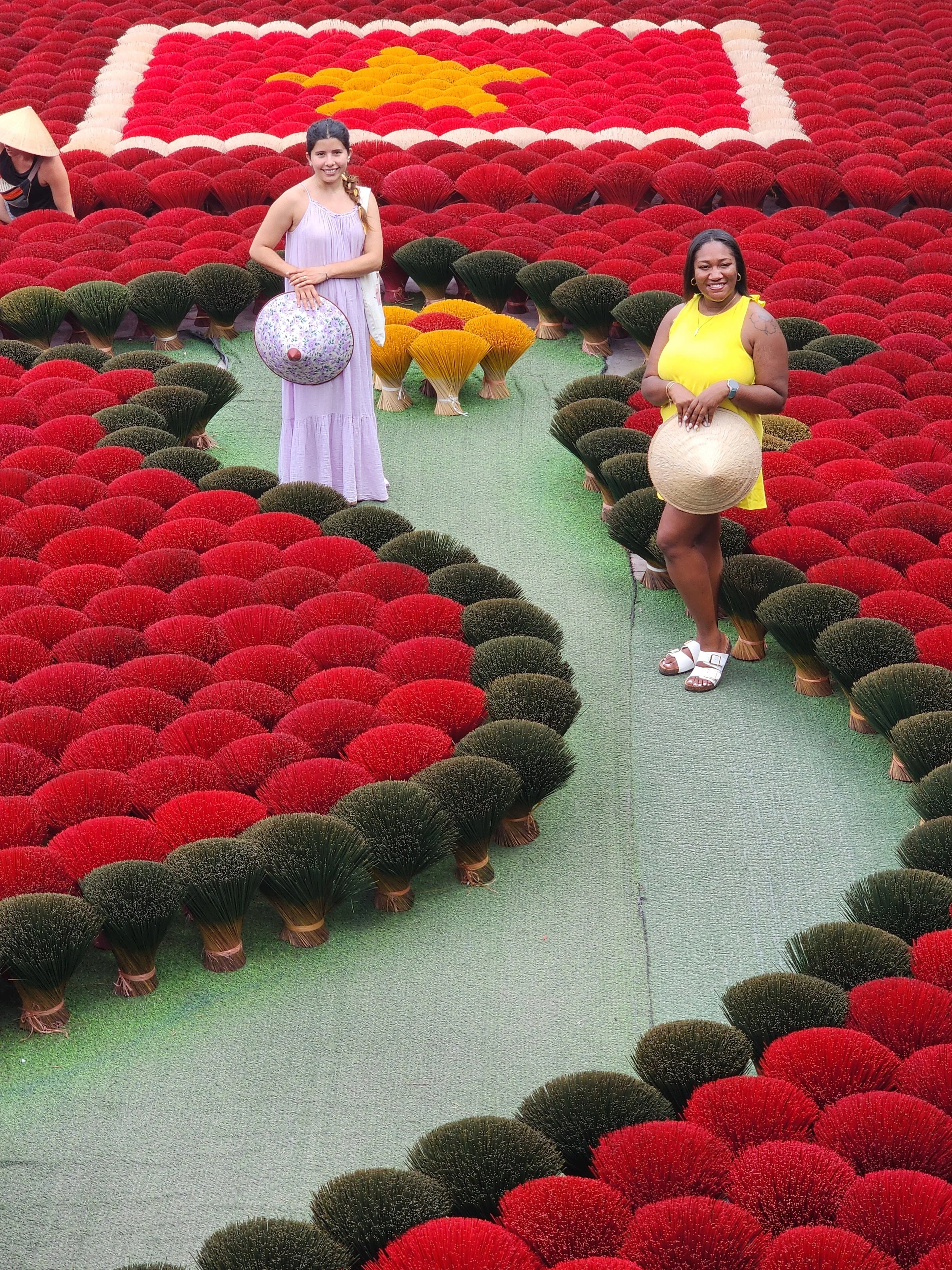 Two women standing among red and green flowers arranged in a pattern, with one woman in a light purple dress holding a round bag and the other in a yellow dress holding a straw hat, in an outdoor flower garden.