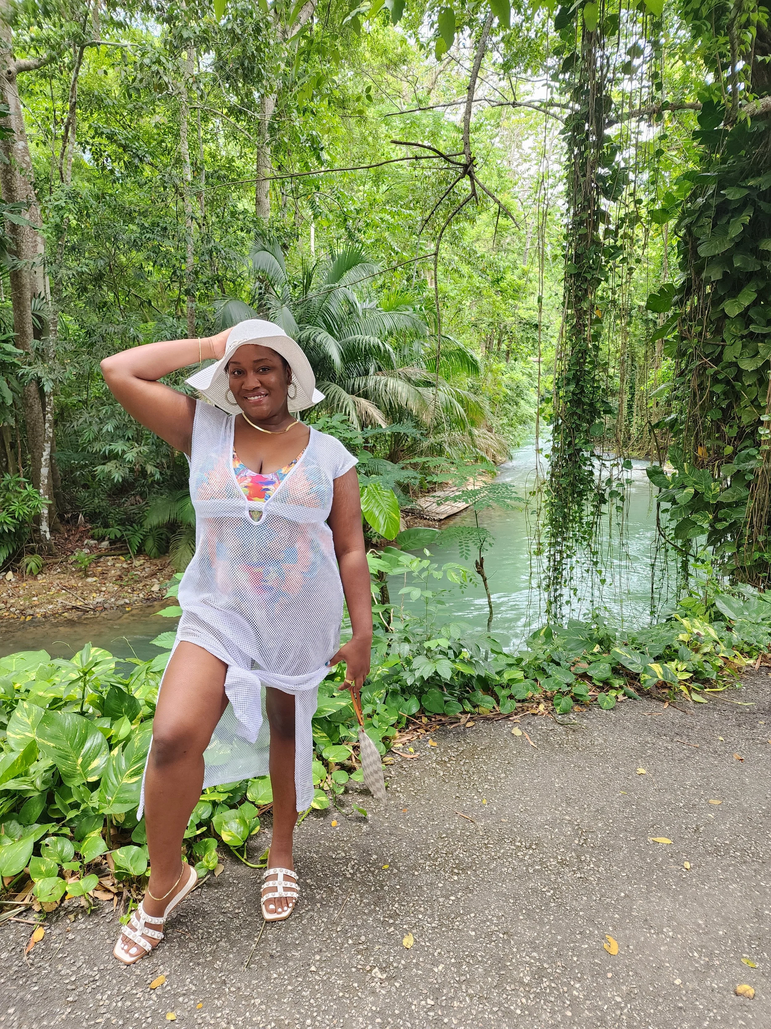 A woman wearing a white hat and flowy cover-up dress stands on a nature trail near a river, surrounded by lush green tropical plants and trees.