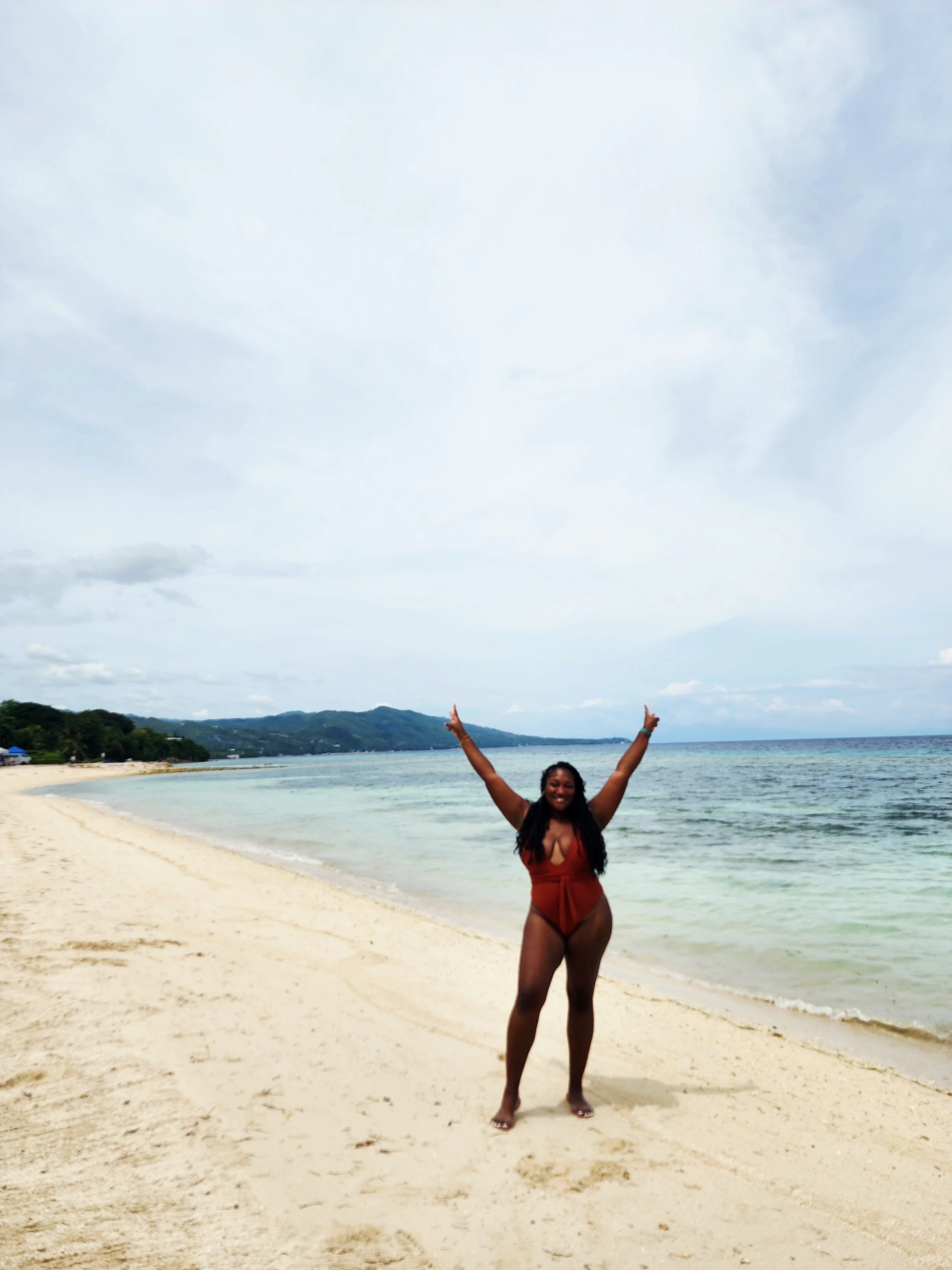 A woman in a red swimsuit standing on a sandy beach with arms raised, smiling, near the shoreline with the ocean and hills in the background.