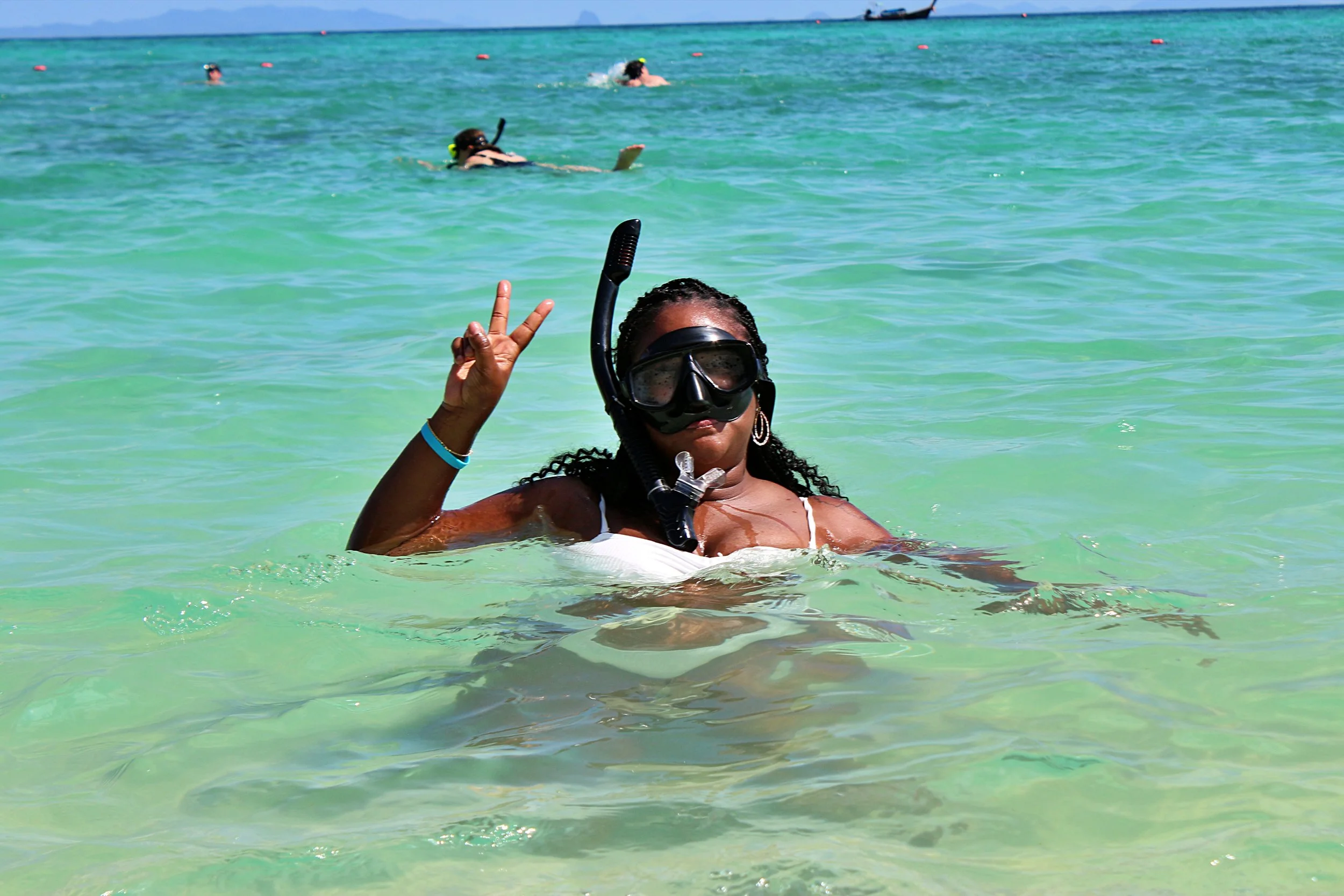 A woman in a white swimsuit wearing a diving mask and snorkel making a peace sign in the ocean, with other swimmers in the background.
