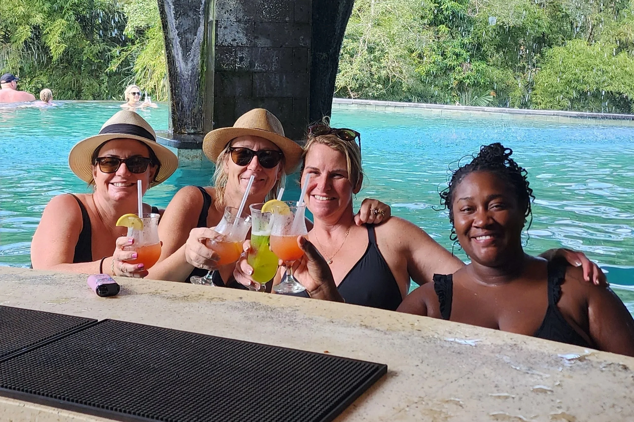 Four women in swimsuits and hats enjoying drinks at a poolside bar, smiling at the camera with a pool and lush green trees in the background.