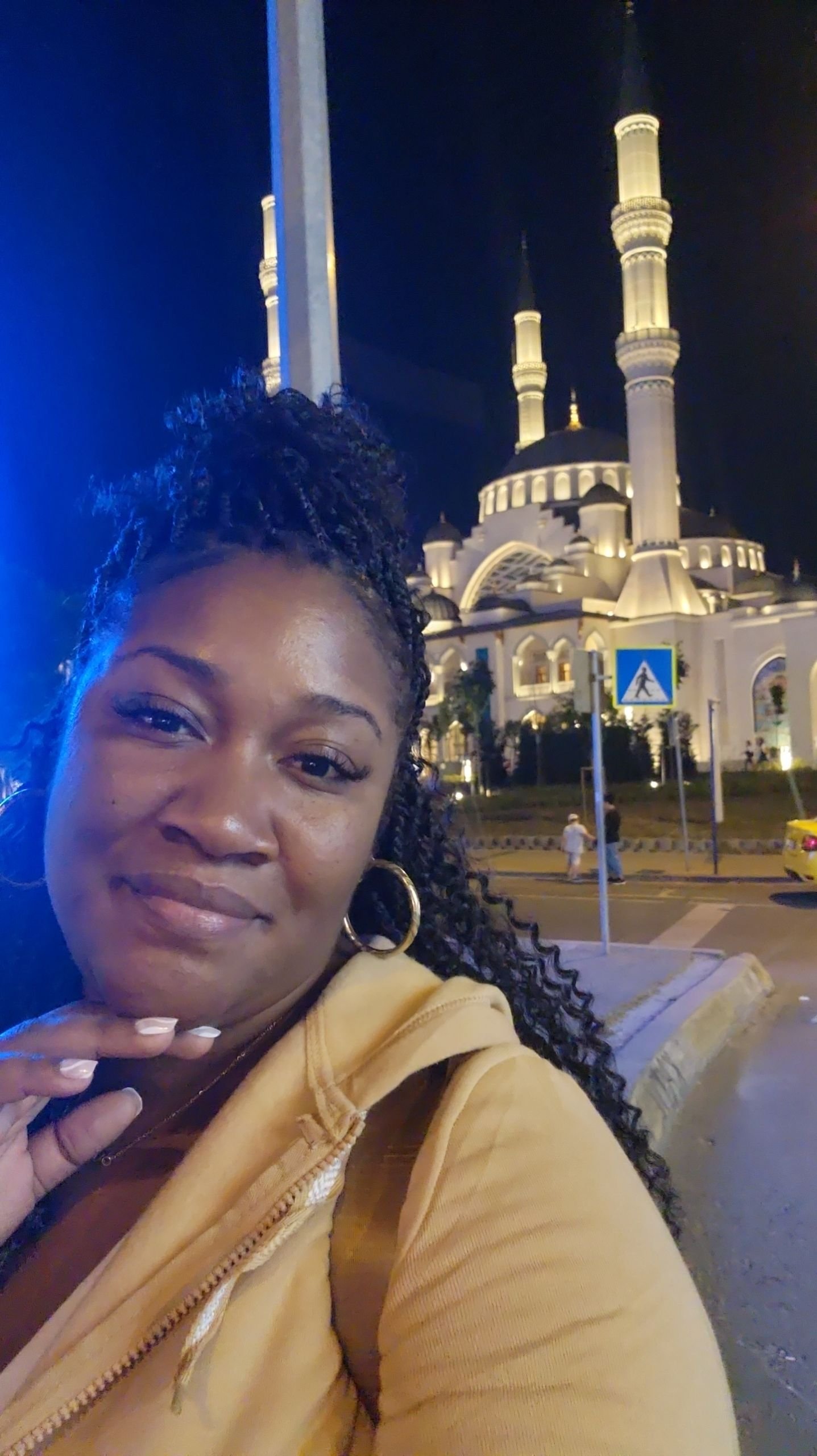 A woman with curly hair and gold hoop earrings taking a selfie in front of an illuminated mosque at night in Turkey
