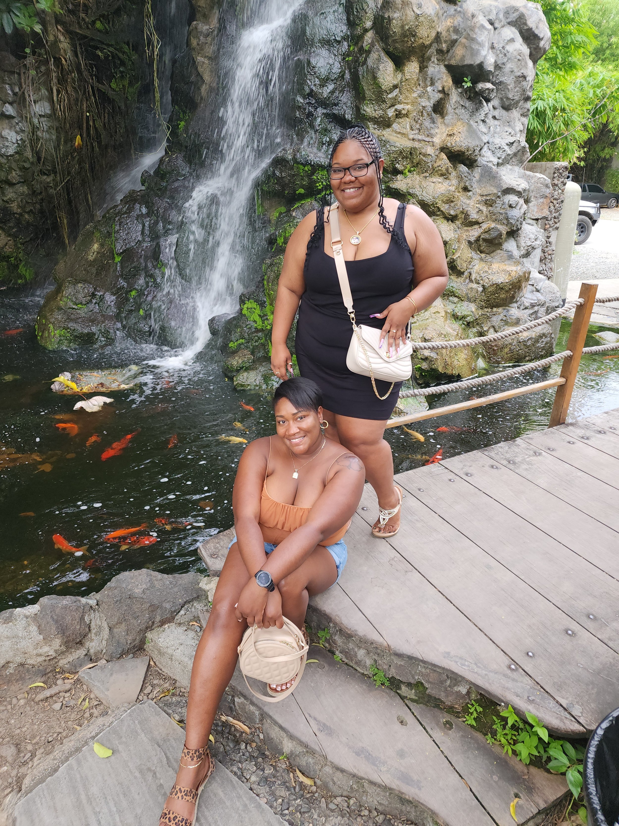 Two women posing near a pond with a waterfall in the background. One is sitting on a rock and the other standing, both smiling. The pond has koi fish swimming, and there is a wooden platform and railing nearby.