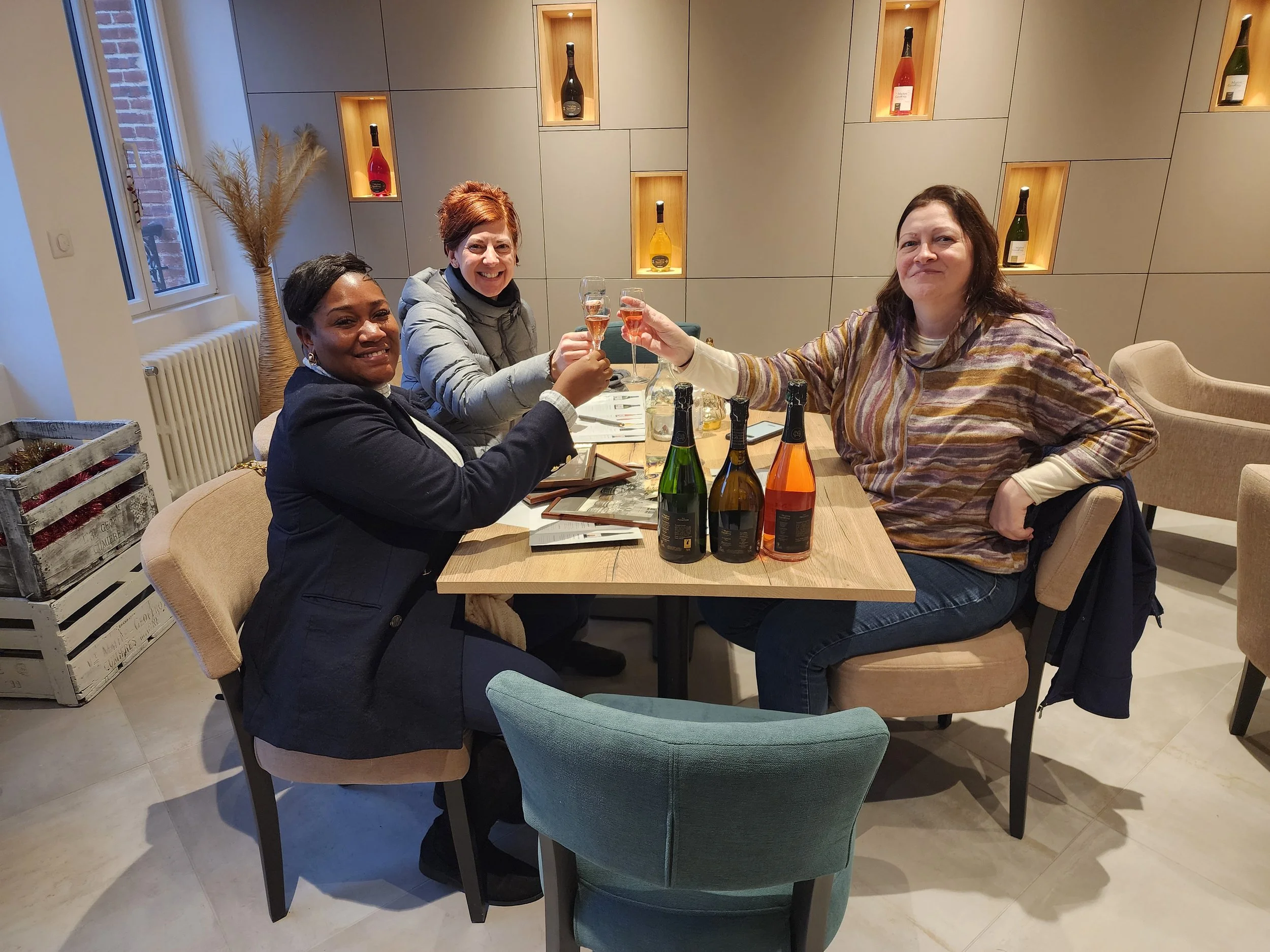 Three women sitting around a table, raising glasses for a toast, with bottles of sparkling wine on the table, in a modern restaurant or cafe.