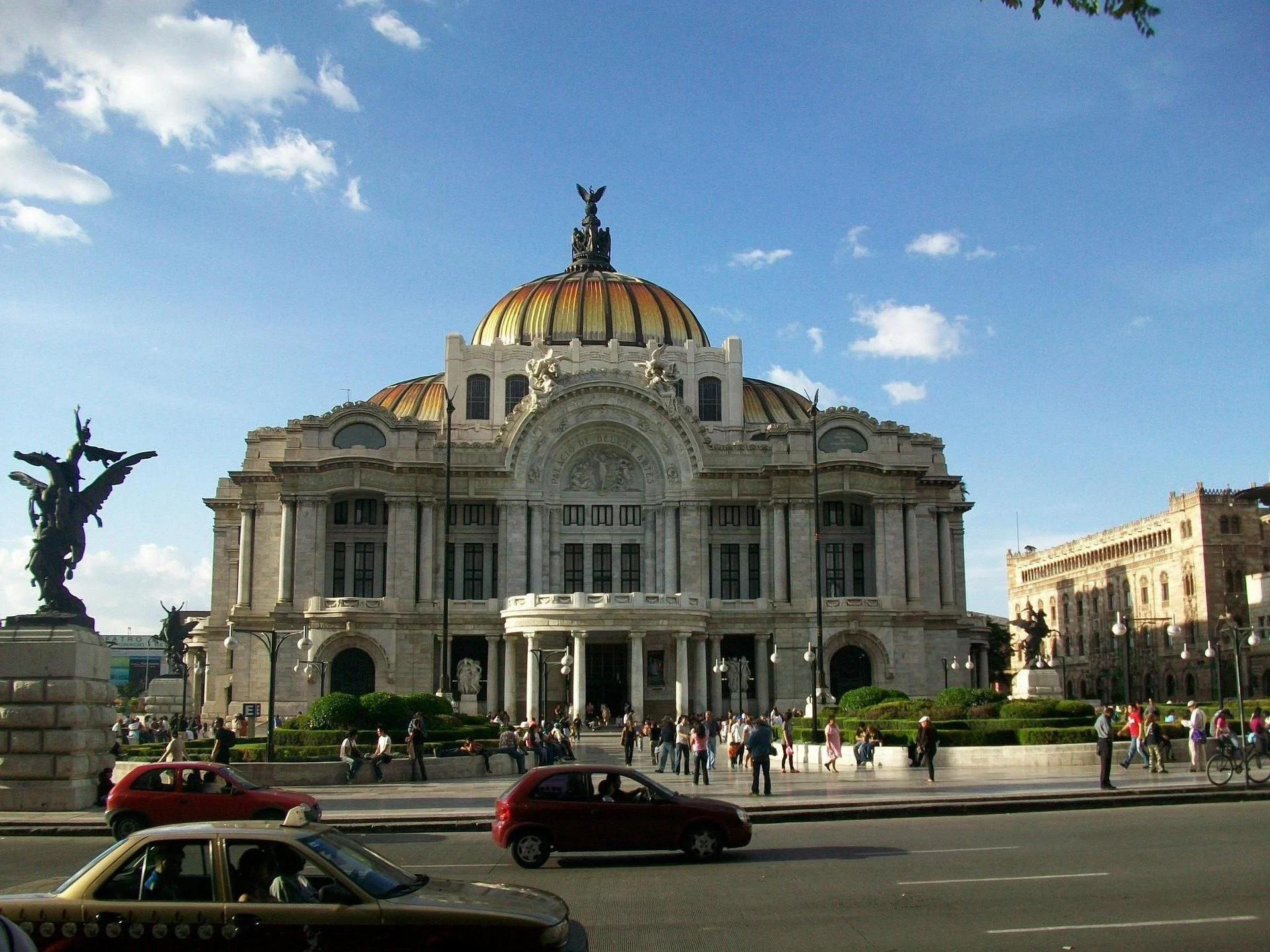The Palace of Bellas Artes in Mexico City with a gold-domed roof, marble facade, and sculptures. People walk in front, and cars drive by on the street.