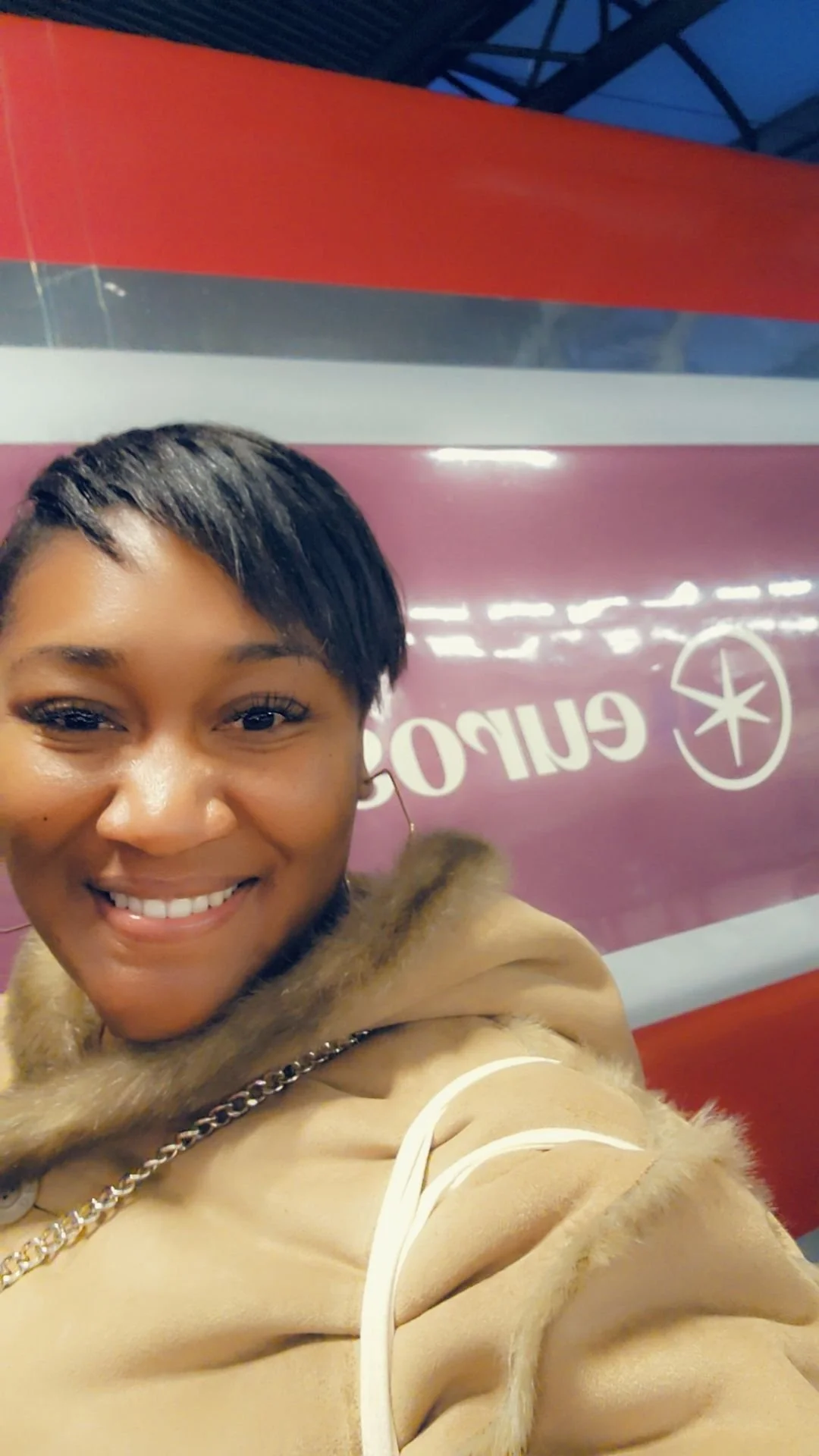 A smiling woman taking a selfie in front of a purple sign with white text and star logo at an airport or train station in Paris