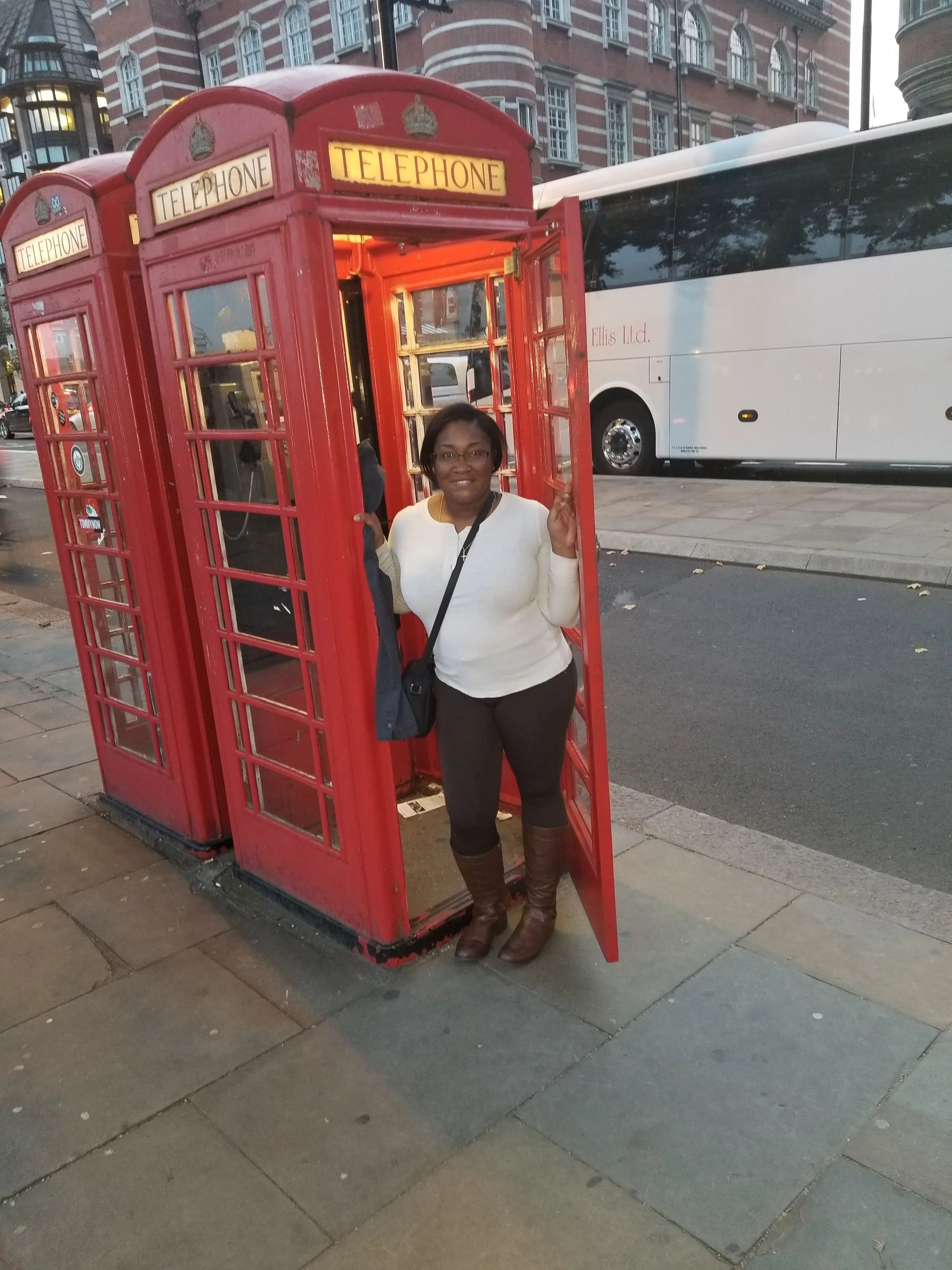 A woman standing inside a classic red British telephone booth on a city sidewalk, dressed in a white top and brown boots, with a large bus in the background.