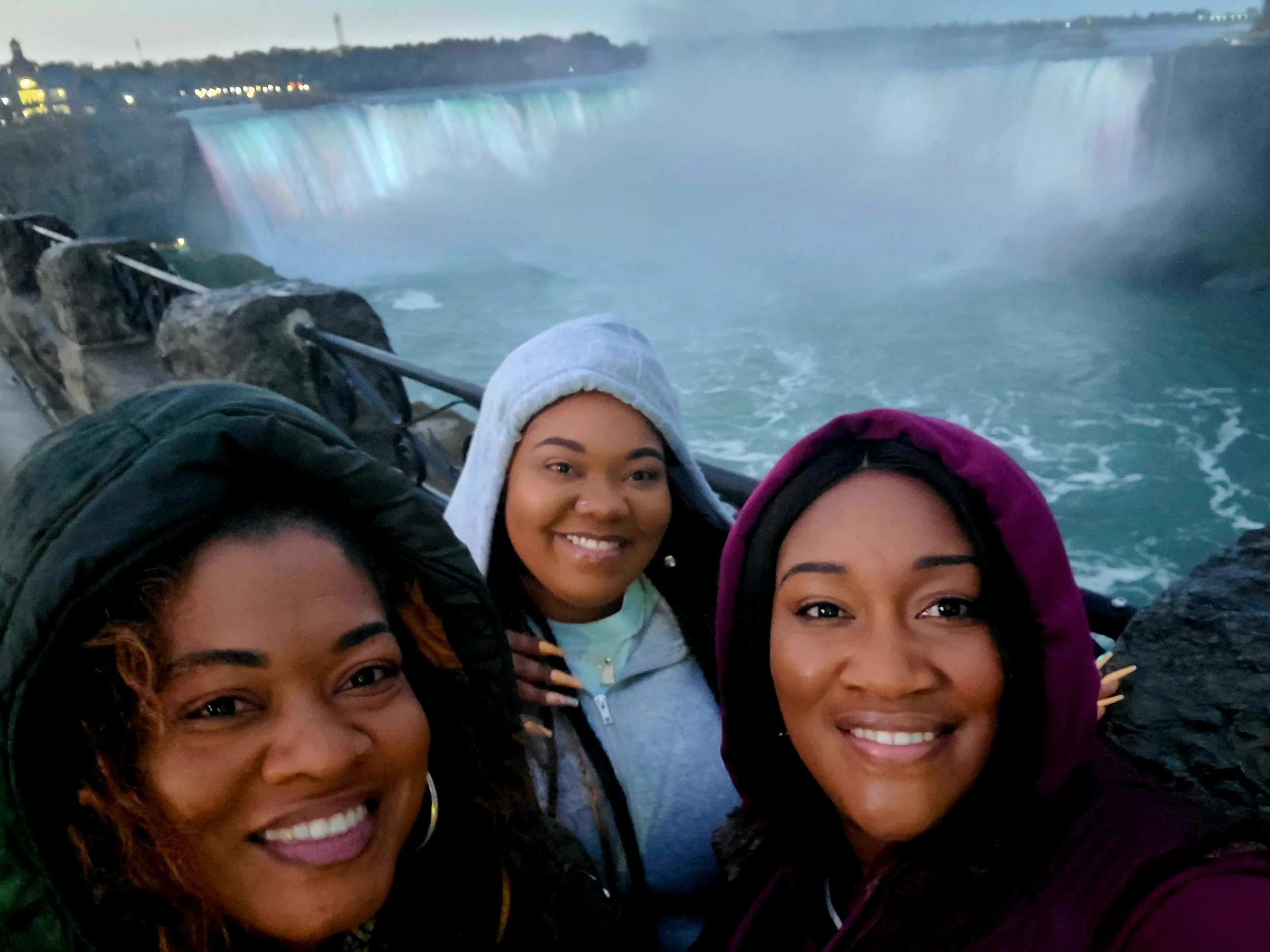Three women taking a selfie at Niagara Falls during dusk, with the waterfall and rainbow mist in the background.