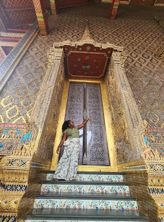 A woman standing on decorated steps in front of an ornate gold and red door inside a temple, pointing towards it.