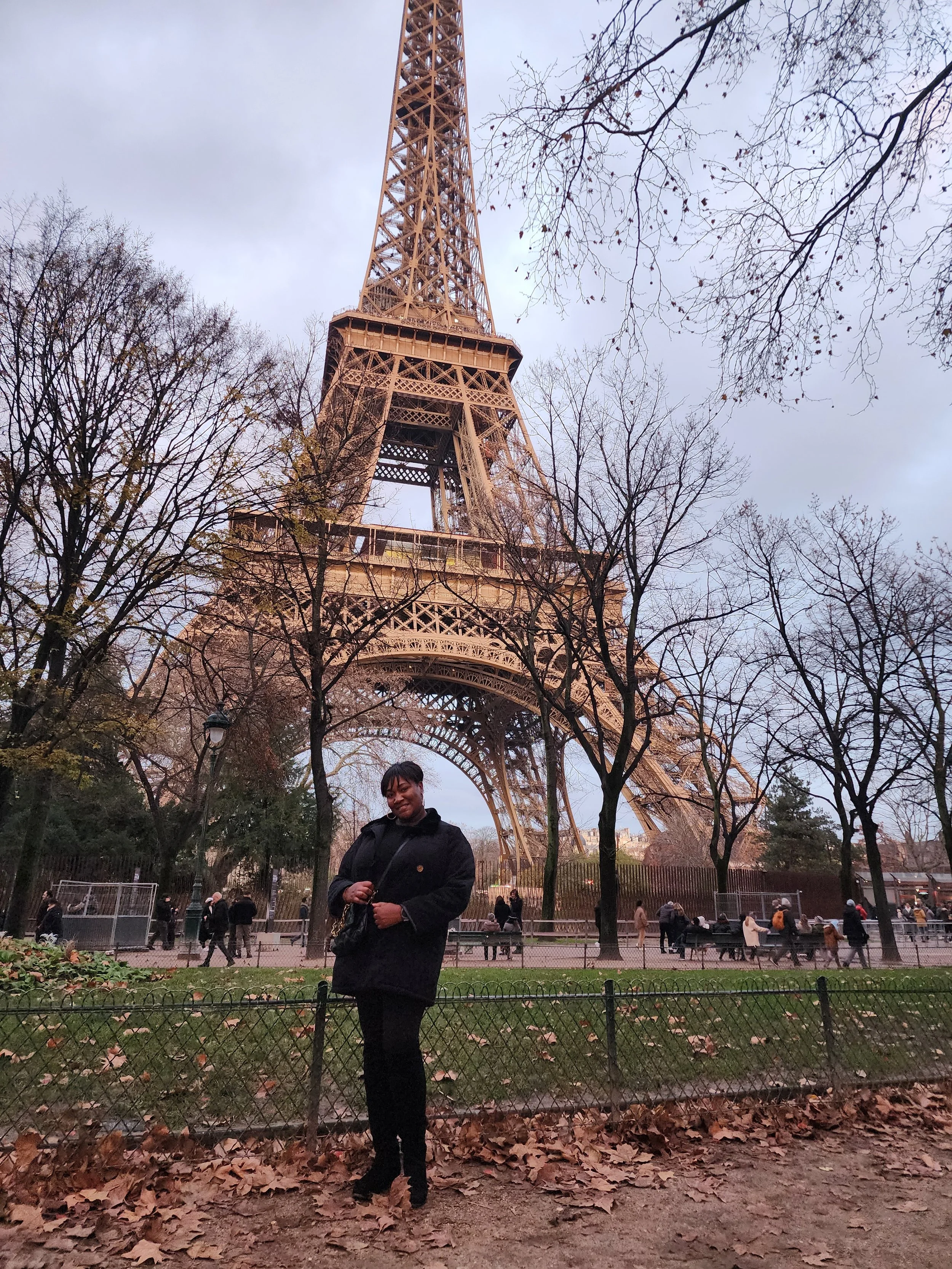 A woman standing in front of the Eiffel Tower in Paris, France, during autumn, with bare trees and fallen leaves on the ground.