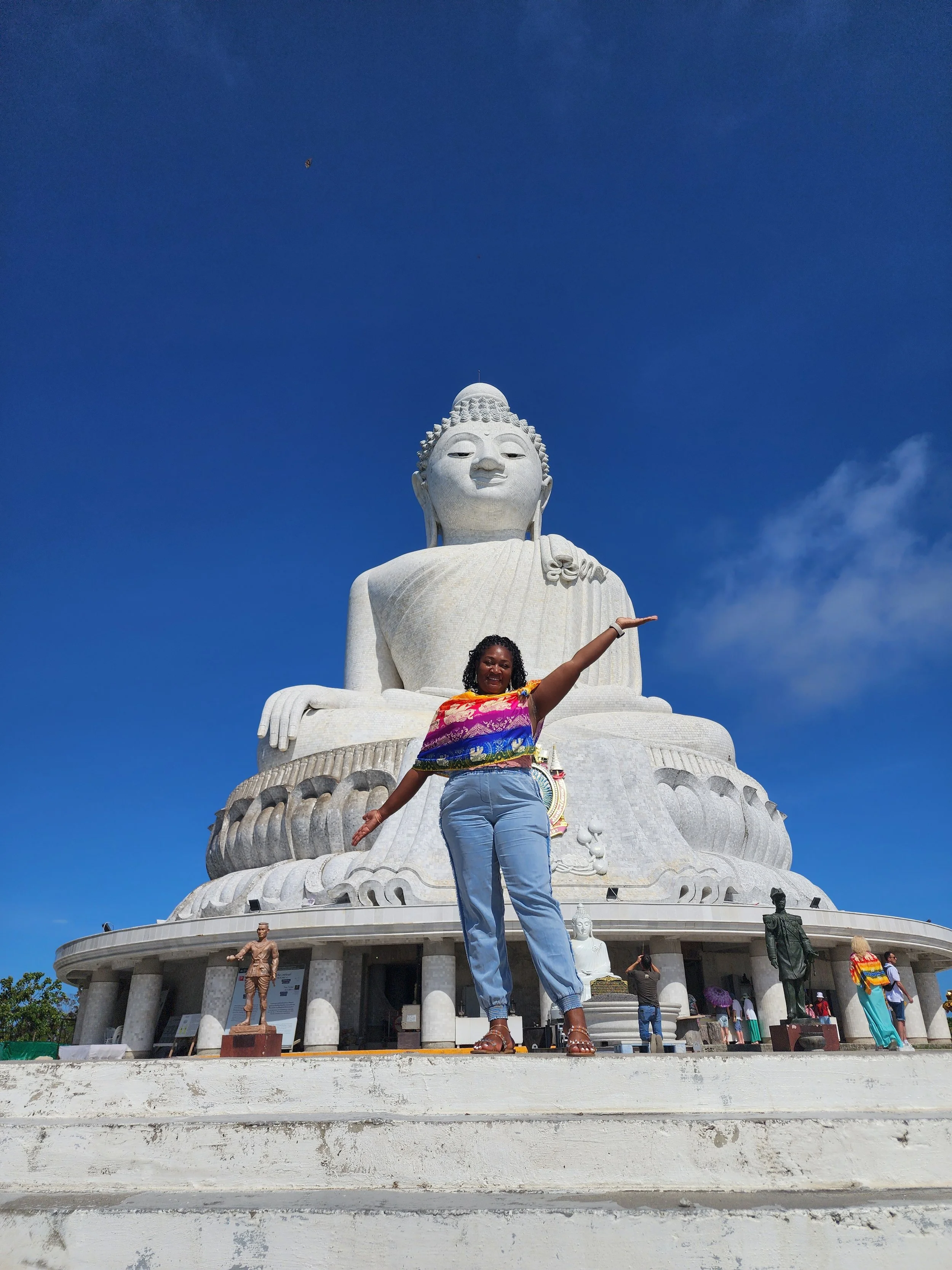 A woman standing in front of a large white Buddha statue in Phuket Thailand, posing with her arm extended upwards. The sky is blue with a few clouds, and there are several smaller statues and visitors around at the site.