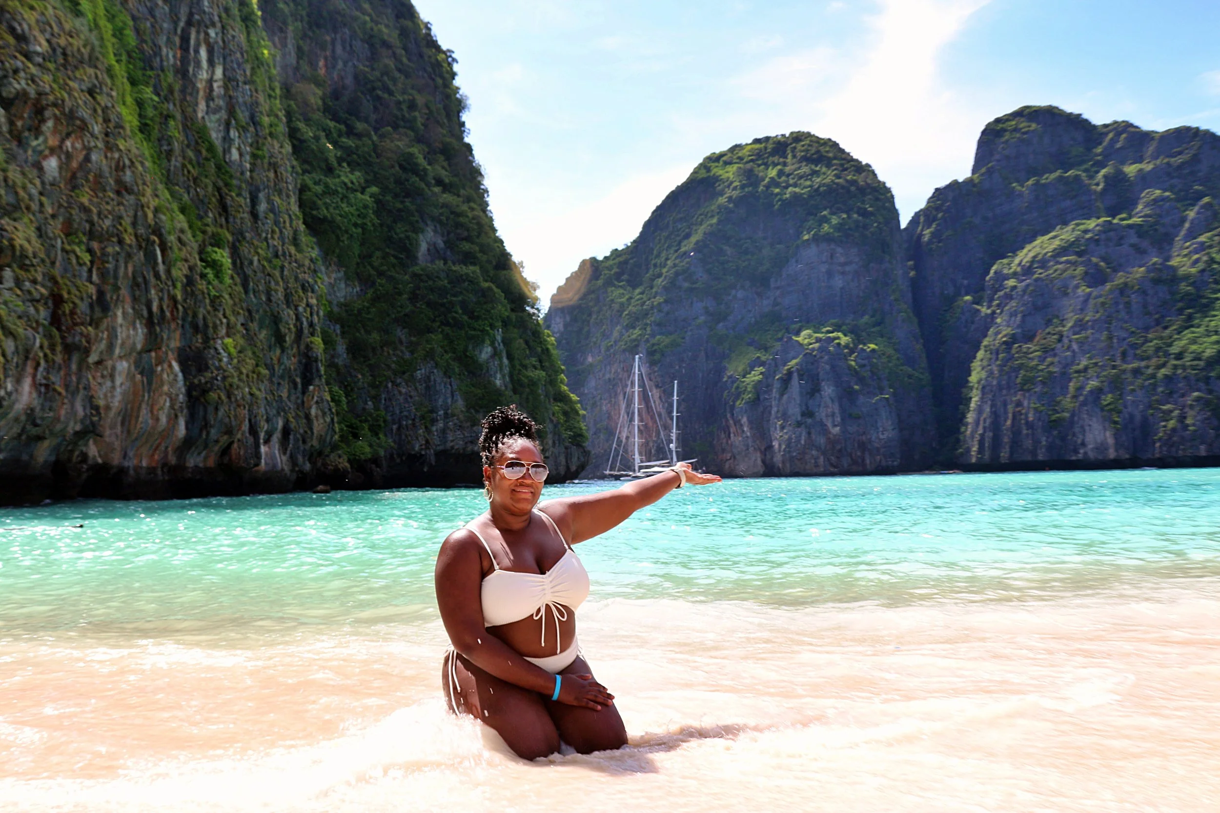 Woman in white bikini kneeling on sandy beach with turquoise water, large rocky cliffs covered in greenery, sailboat in distance, bright sunny sky.