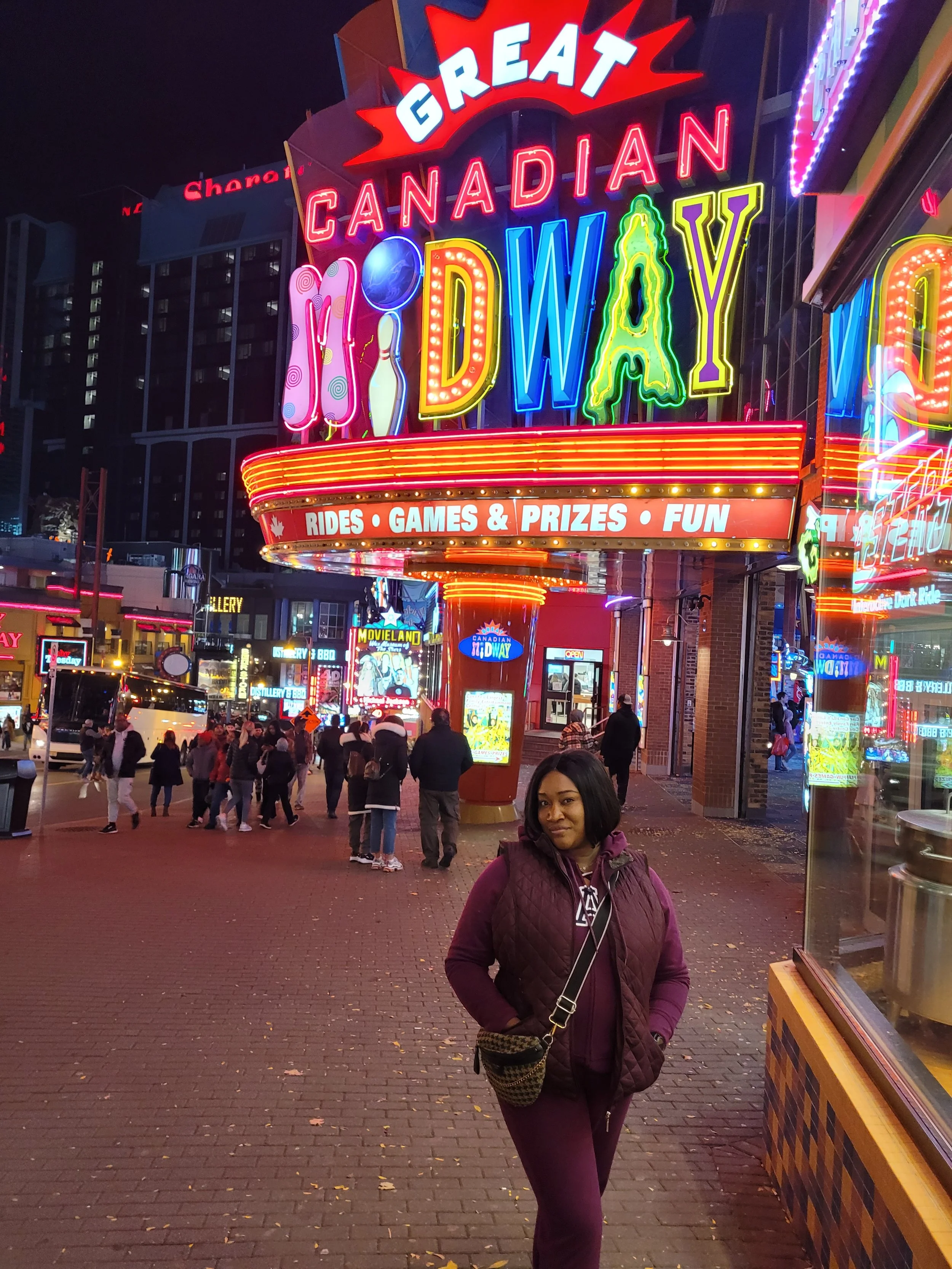 A woman standing in front of the Great Canadian Mideway amusement park at night, with neon lights and a crowd of people in the background.