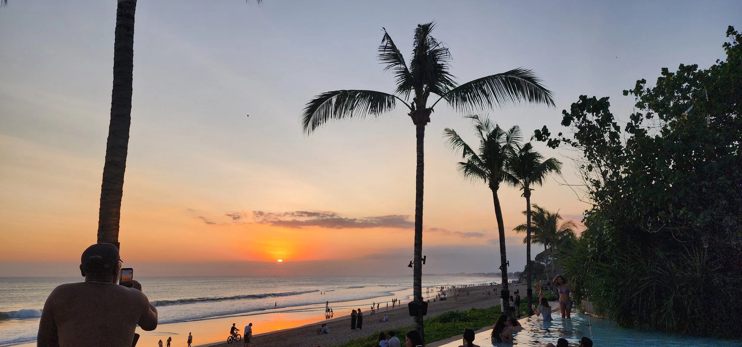 Sunset at the beach with palm trees, people swimming and walking, some taking photos, tropical greenery, and a pool on the right.