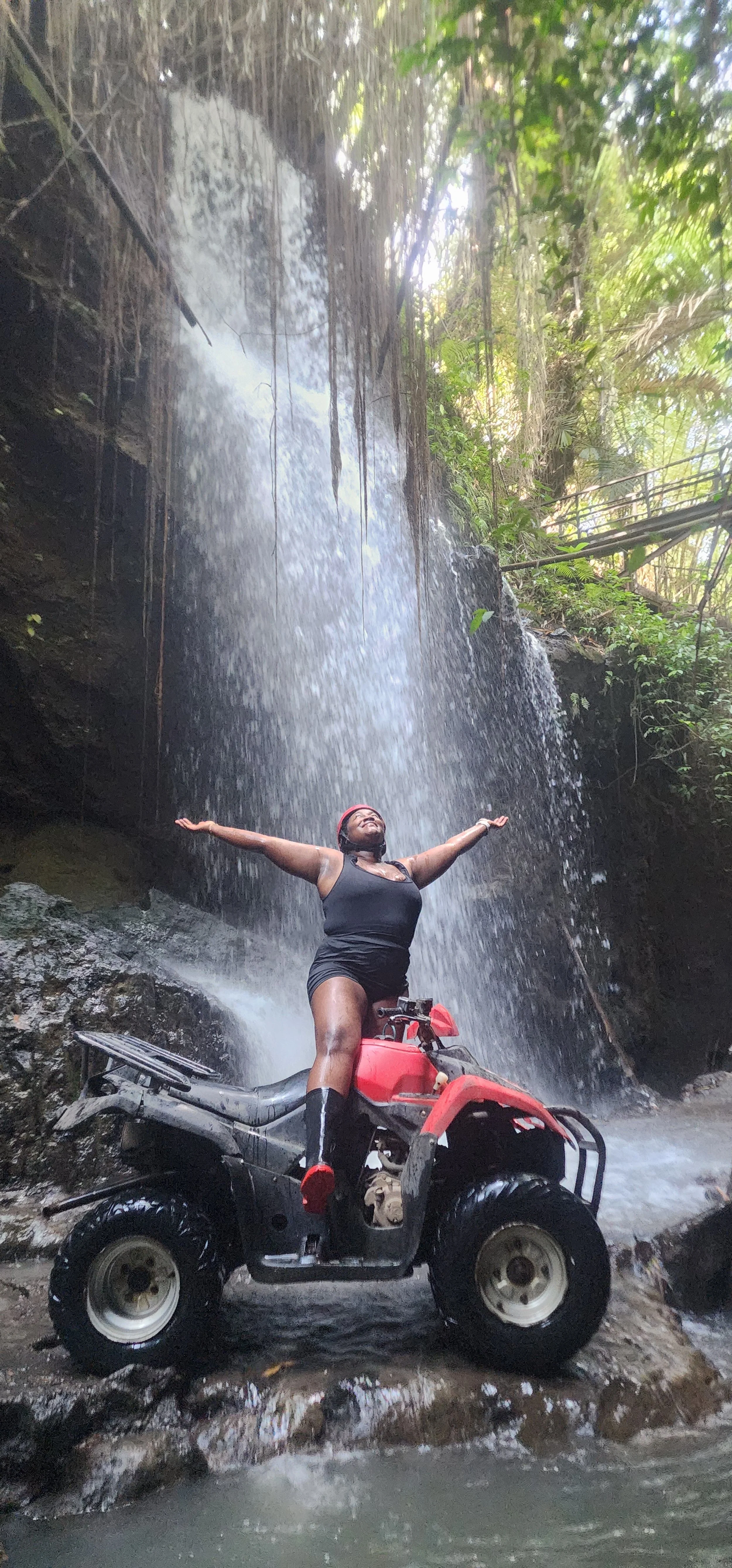 A woman standing on an all-terrain vehicle under a waterfall with arms outstretched in a forest setting.