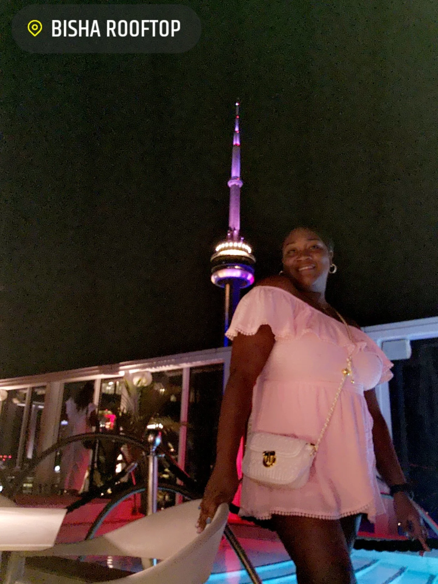 A woman in a pink dress standing on a rooftop at night with the CN Tower lit up in the background.