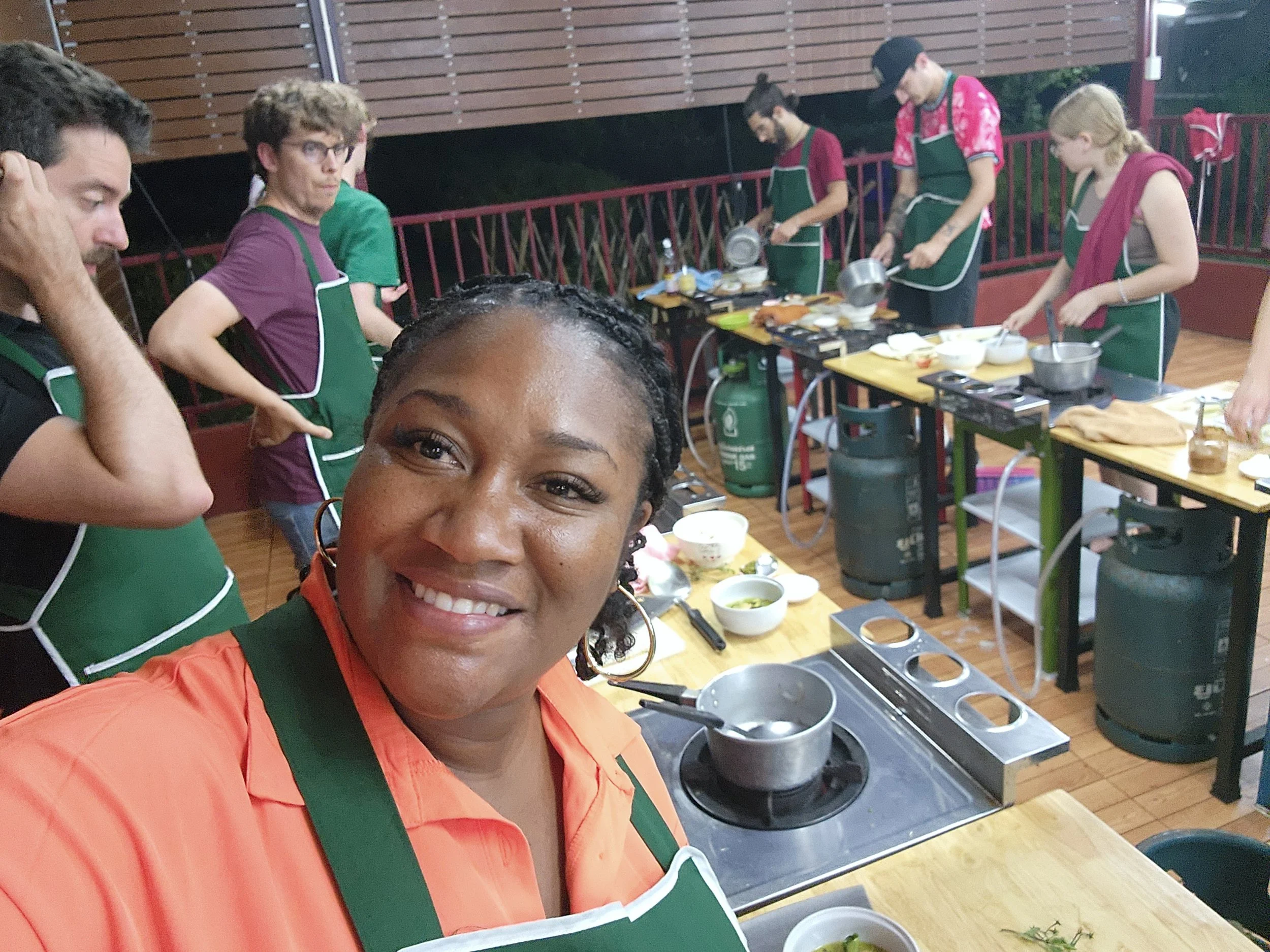 A woman taking a selfie in a cooking class with a group of people preparing food in the background.