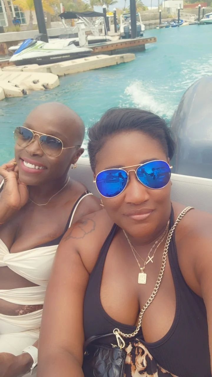 Two women wearing sunglasses taking a selfie on a boat, with water and boats in the background.