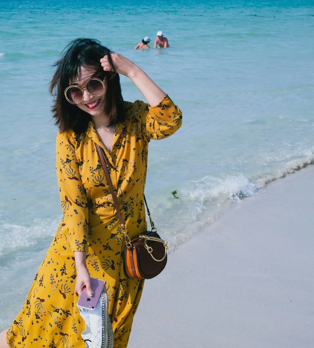 A woman in a yellow floral dress with sunglasses smiling at the beach, holding her phone and a small bag, with the ocean and two people swimming in the background.